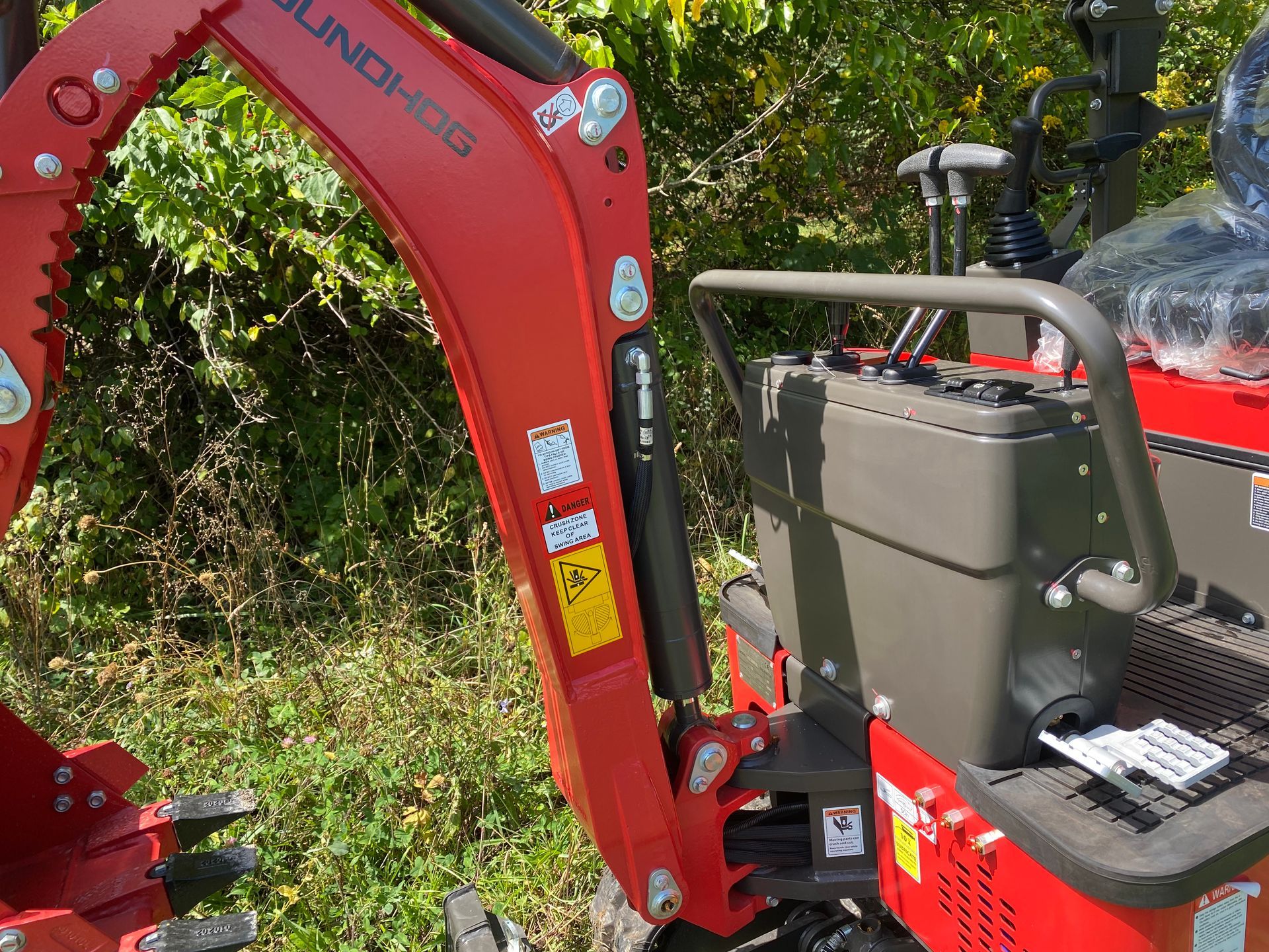 Red mini-excavator with hydraulic arm, near foliage.