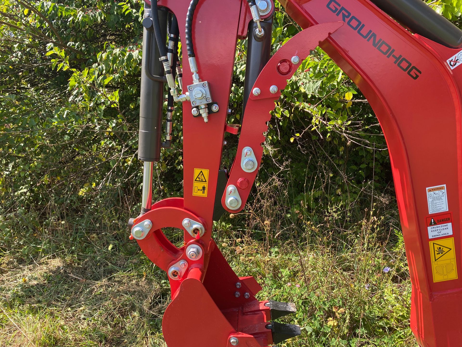 Red Groundhog backhoe arm with hydraulic hoses and bucket. Outdoors, in front of foliage.