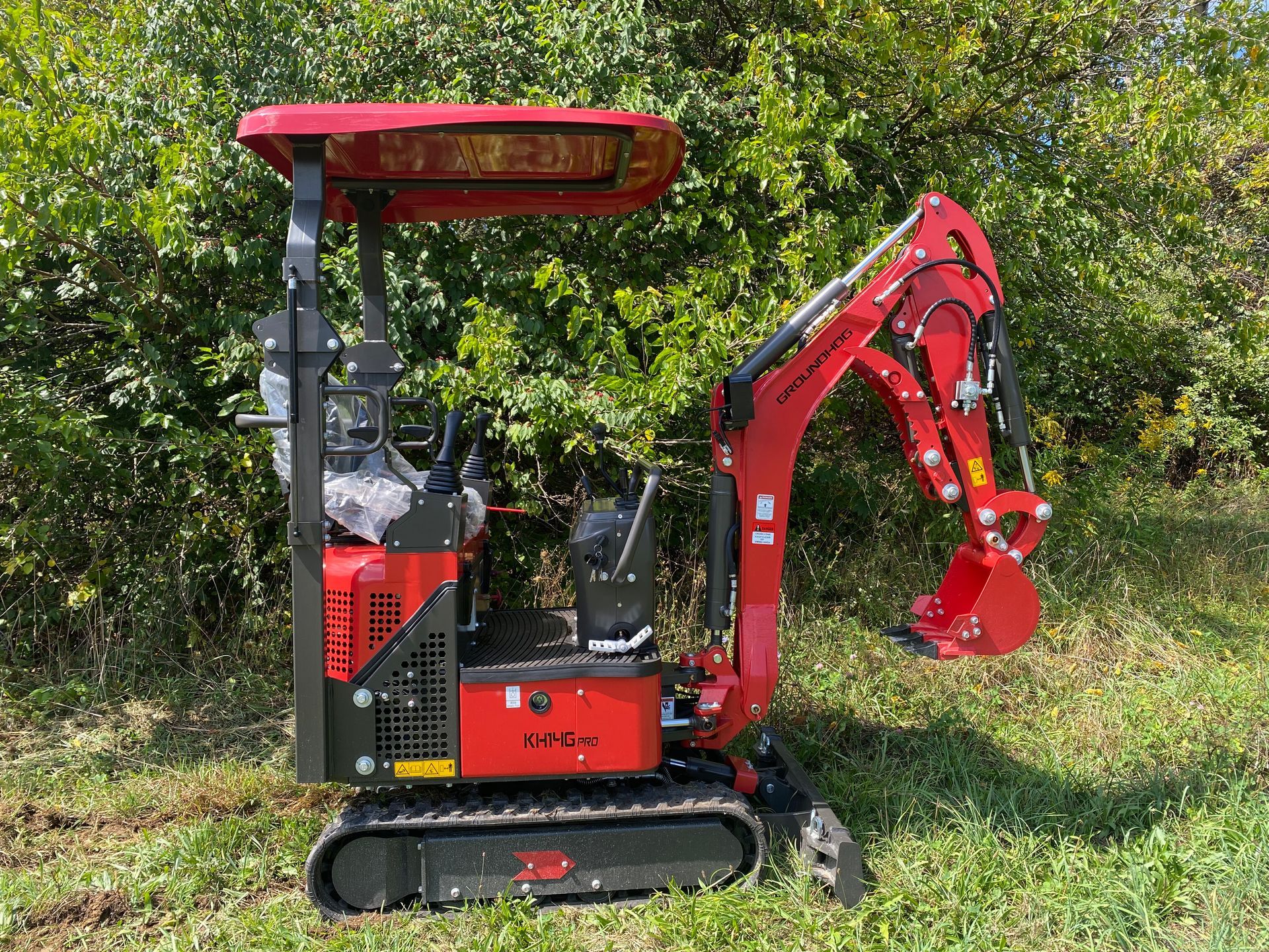 Red mini excavator on tracks in a grassy area with trees in the background.