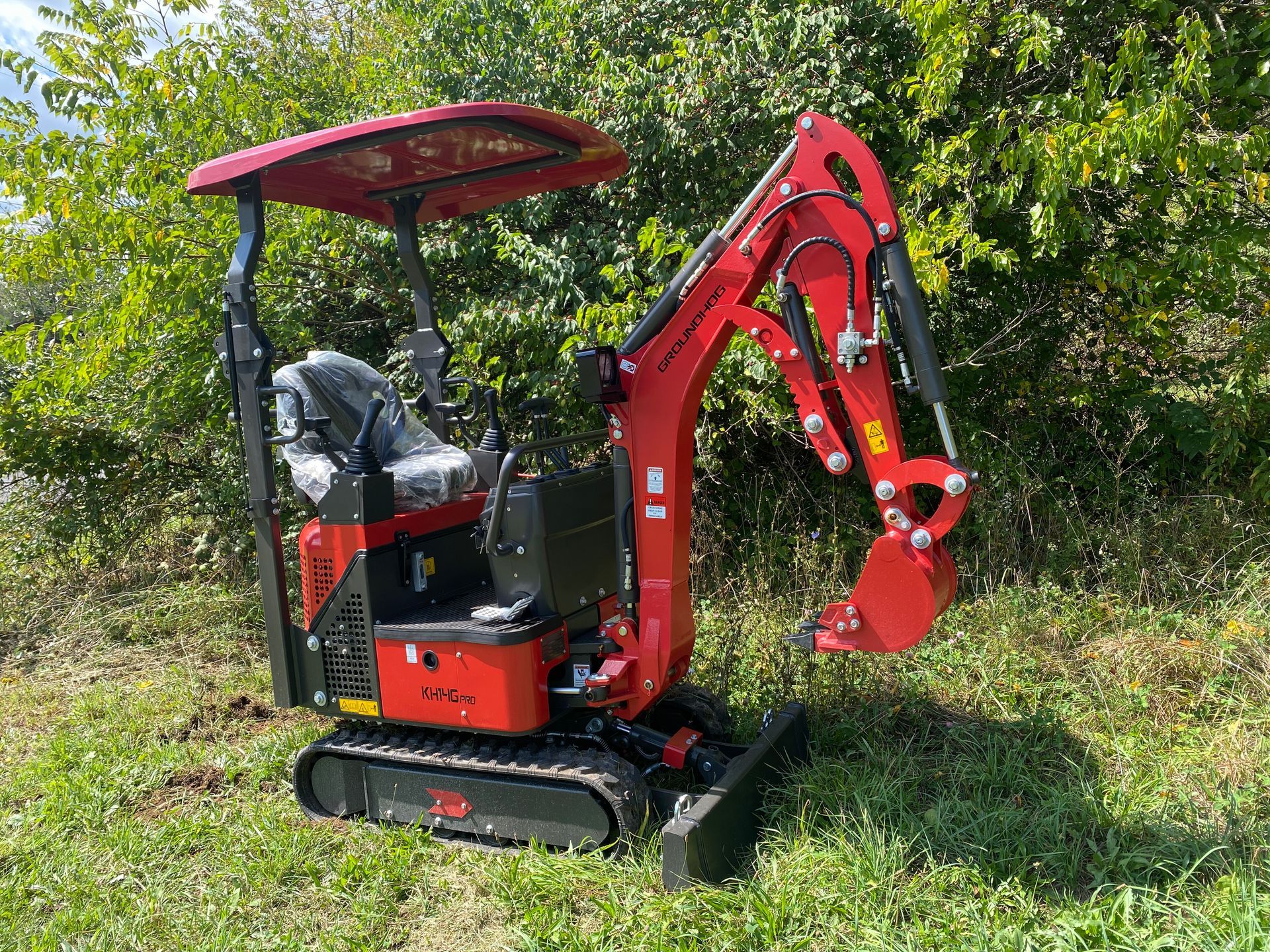 Red mini excavator on grass in front of bushes.