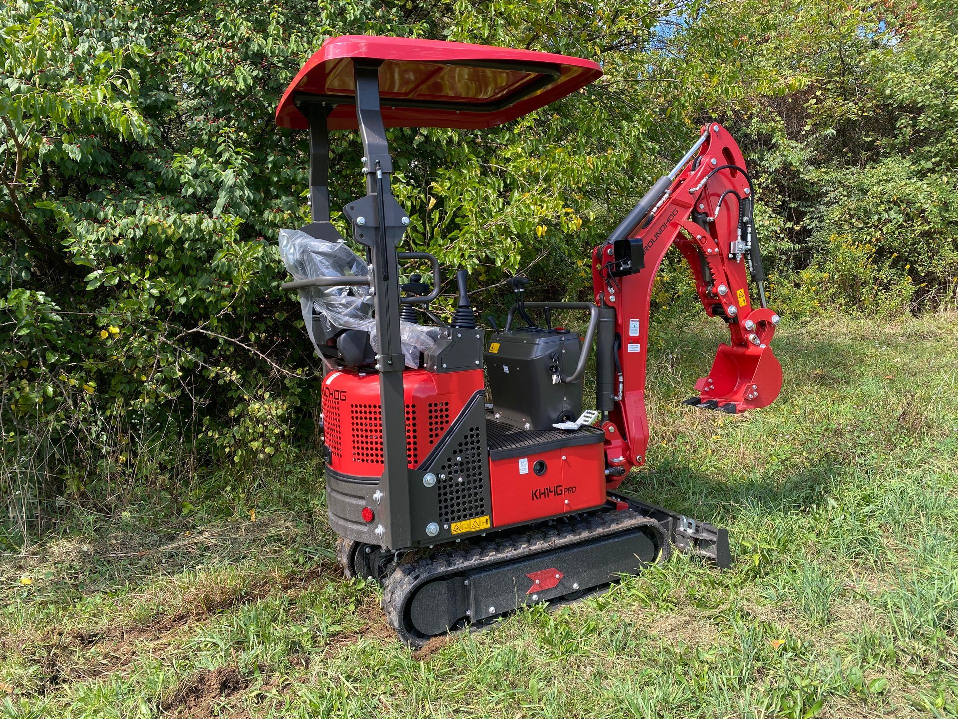 Red mini excavator with canopy in grassy area near trees.