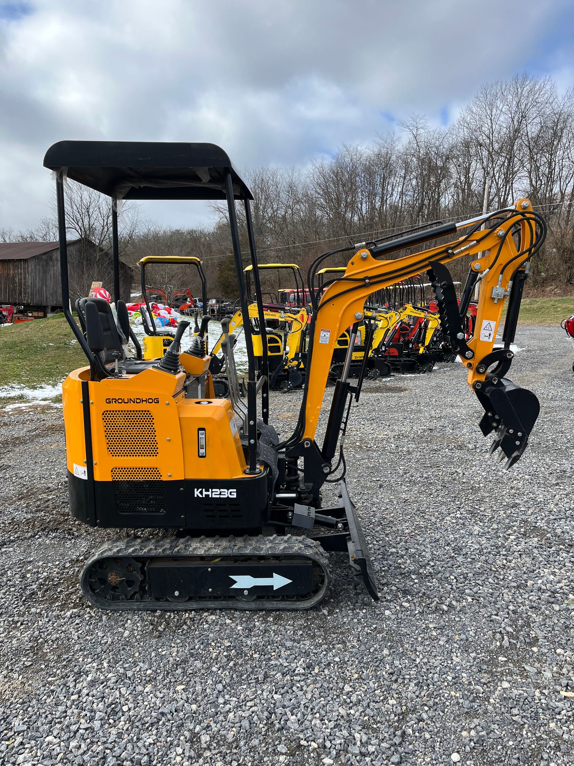 Yellow mini excavator on gravel, outside with trees and other machinery in the background.