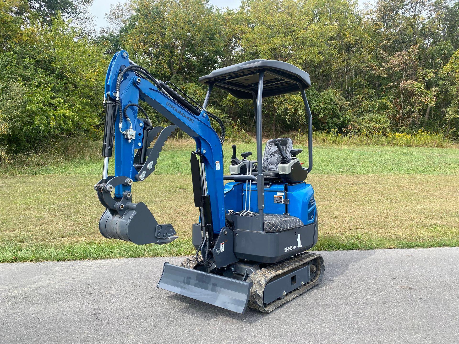 Blue mini excavator on a paved surface, parked with bucket down, in front of trees and grass.