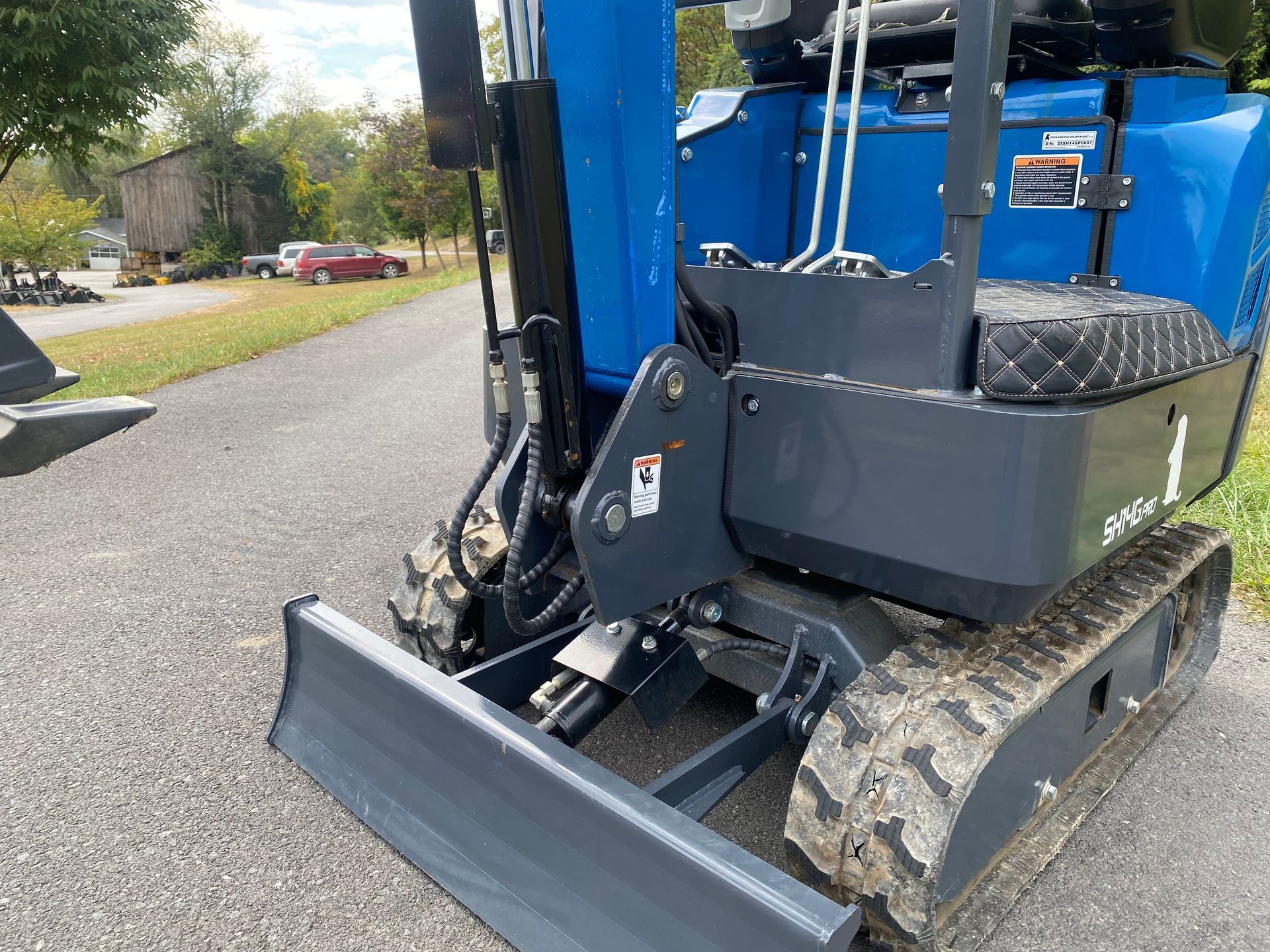 Blue and gray mini excavator on asphalt driveway, with a small building in the background.