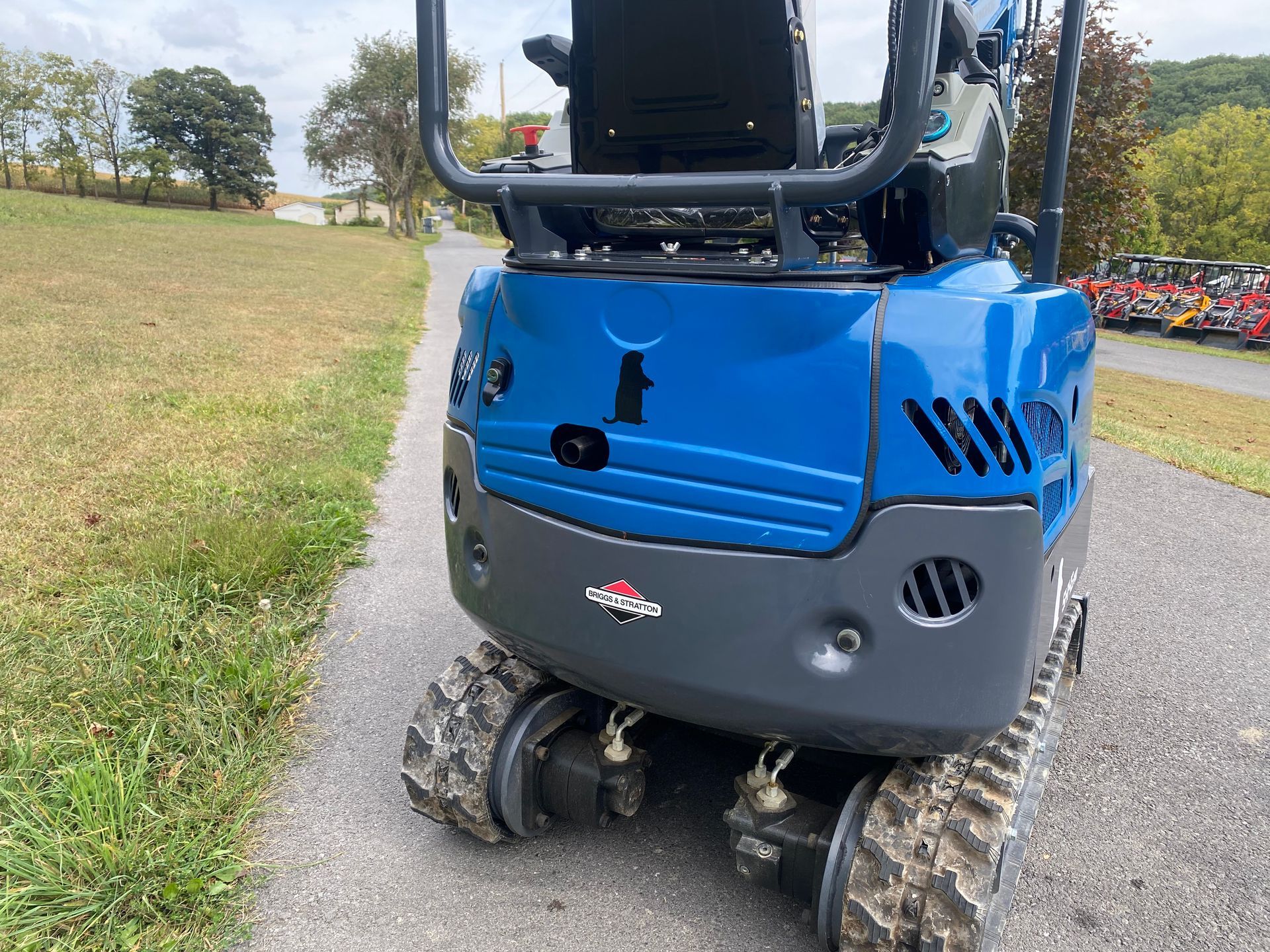 Blue mini excavator on a gray asphalt path with grassy hill, trees, and machinery in the background.