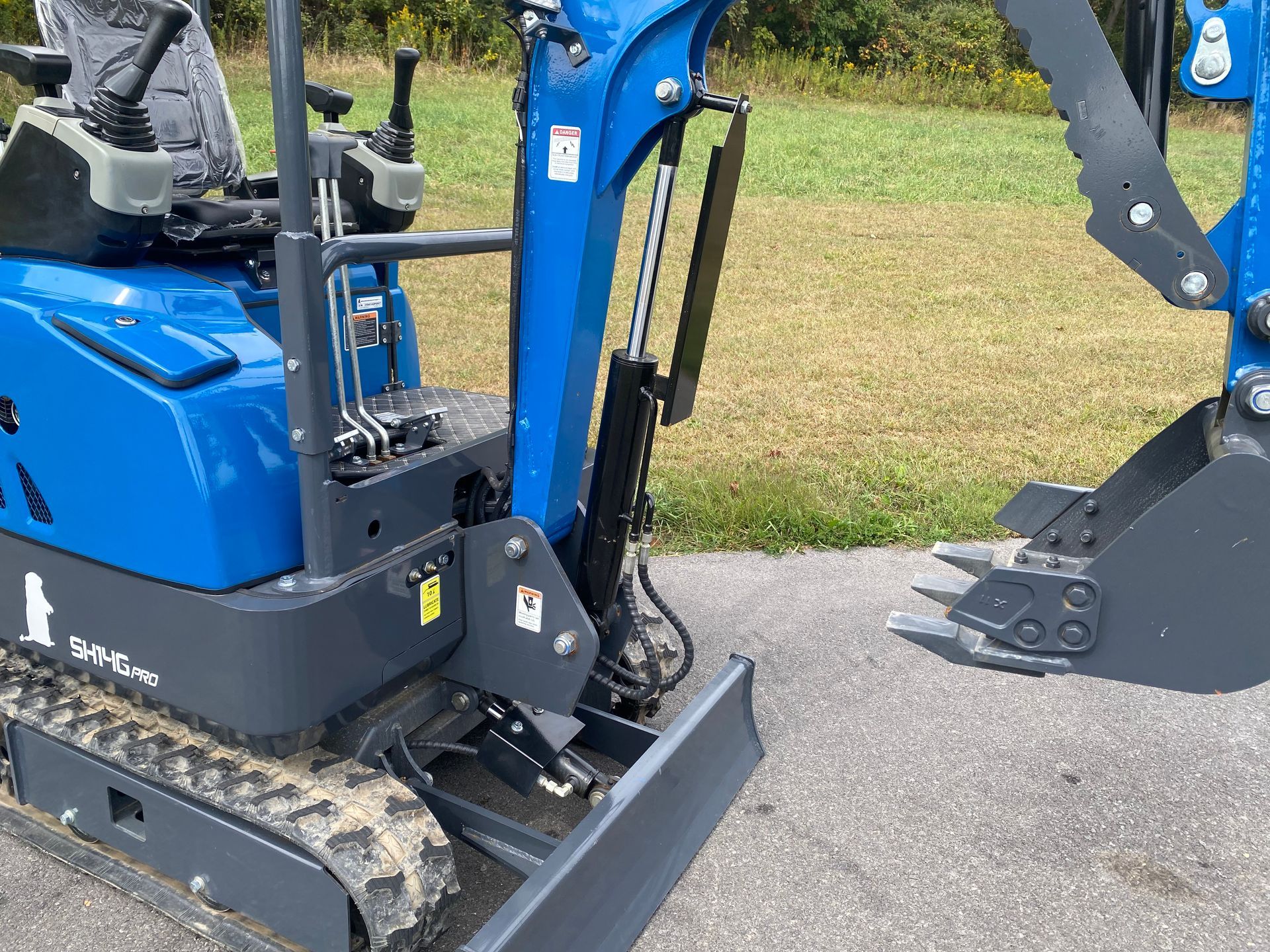 Blue mini excavator on asphalt, bucket and blade in view, green field background.