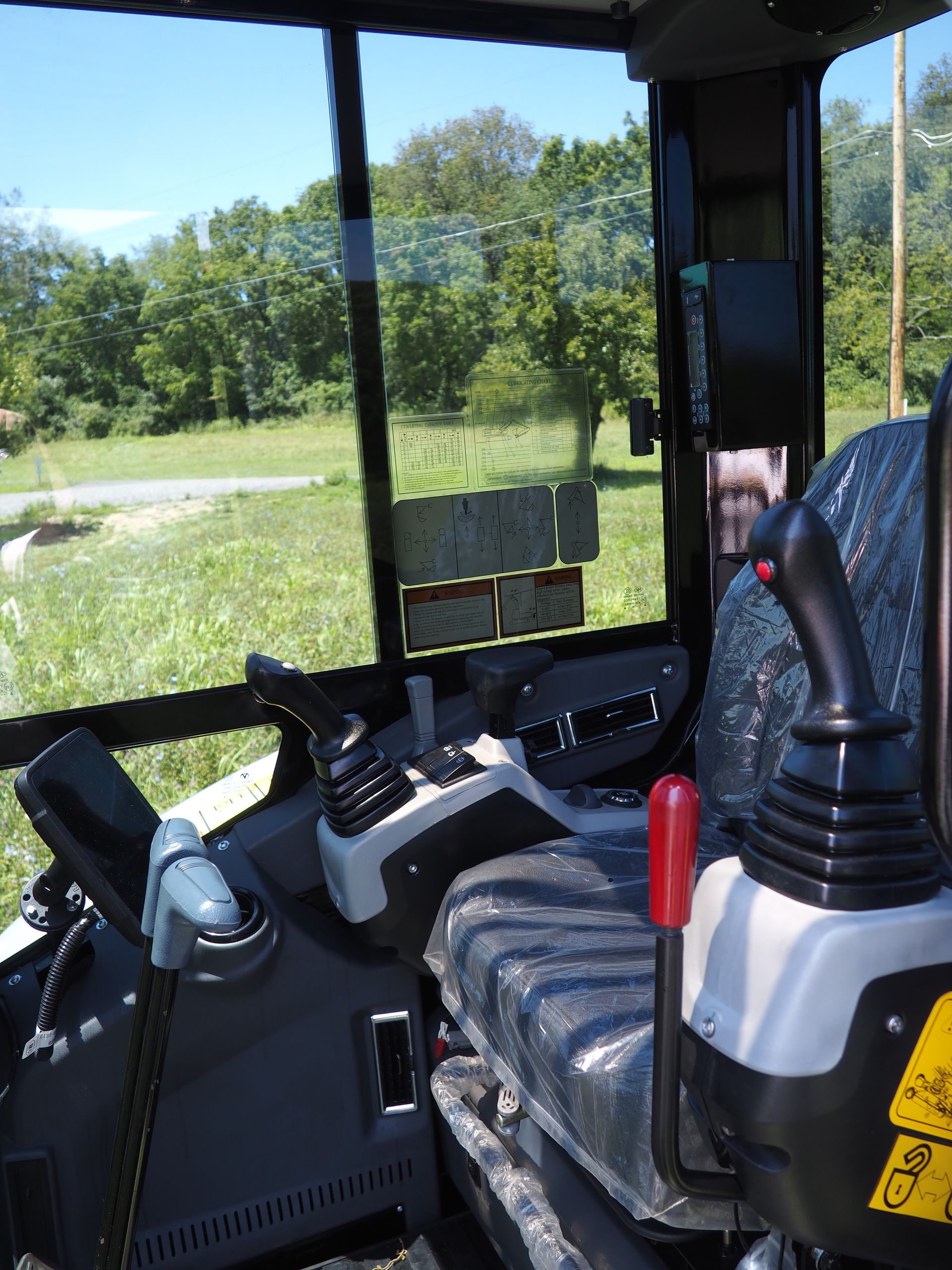 Interior of an excavator cab. Levers, controls, and seat visible. View through the window of trees and grass.