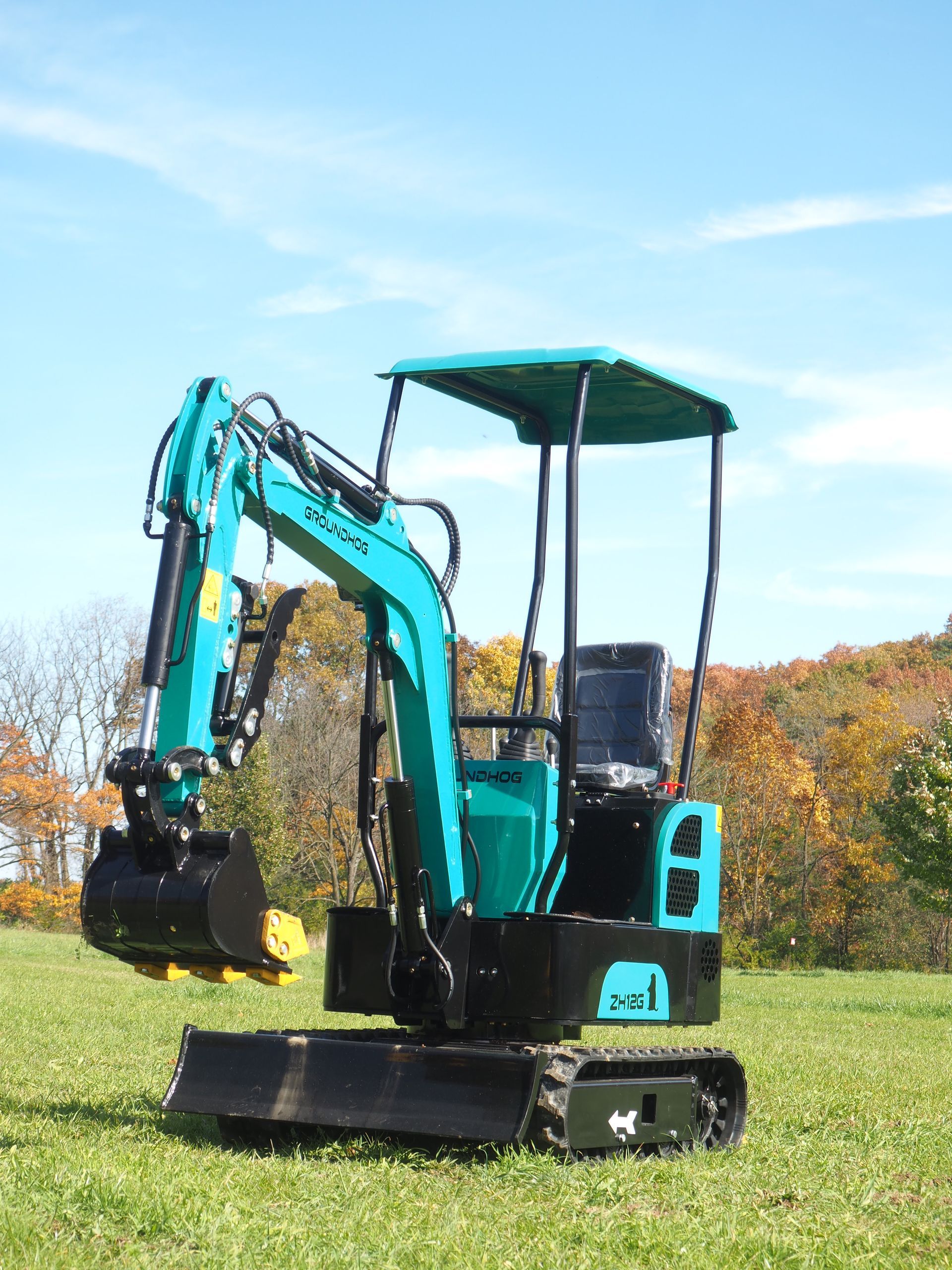 A teal mini-excavator on tracks in a grassy field with autumn trees in the background.