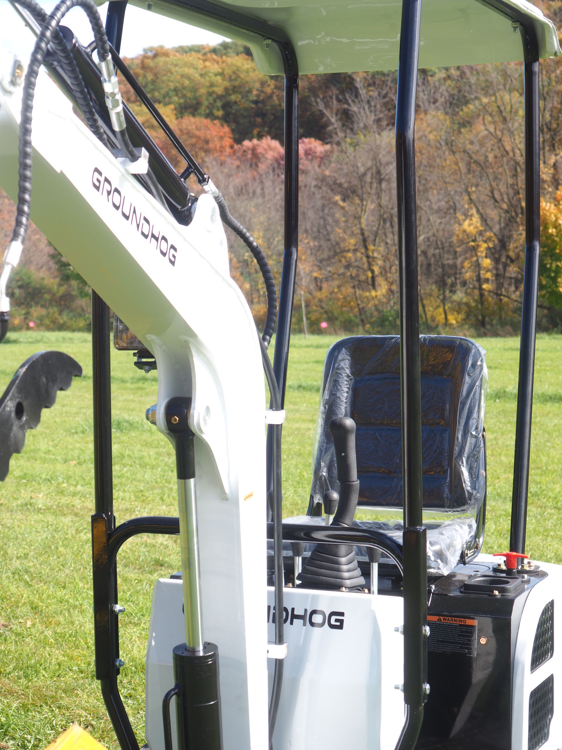 Mini-excavator with roll cage and seat in outdoor setting, autumn trees in background.