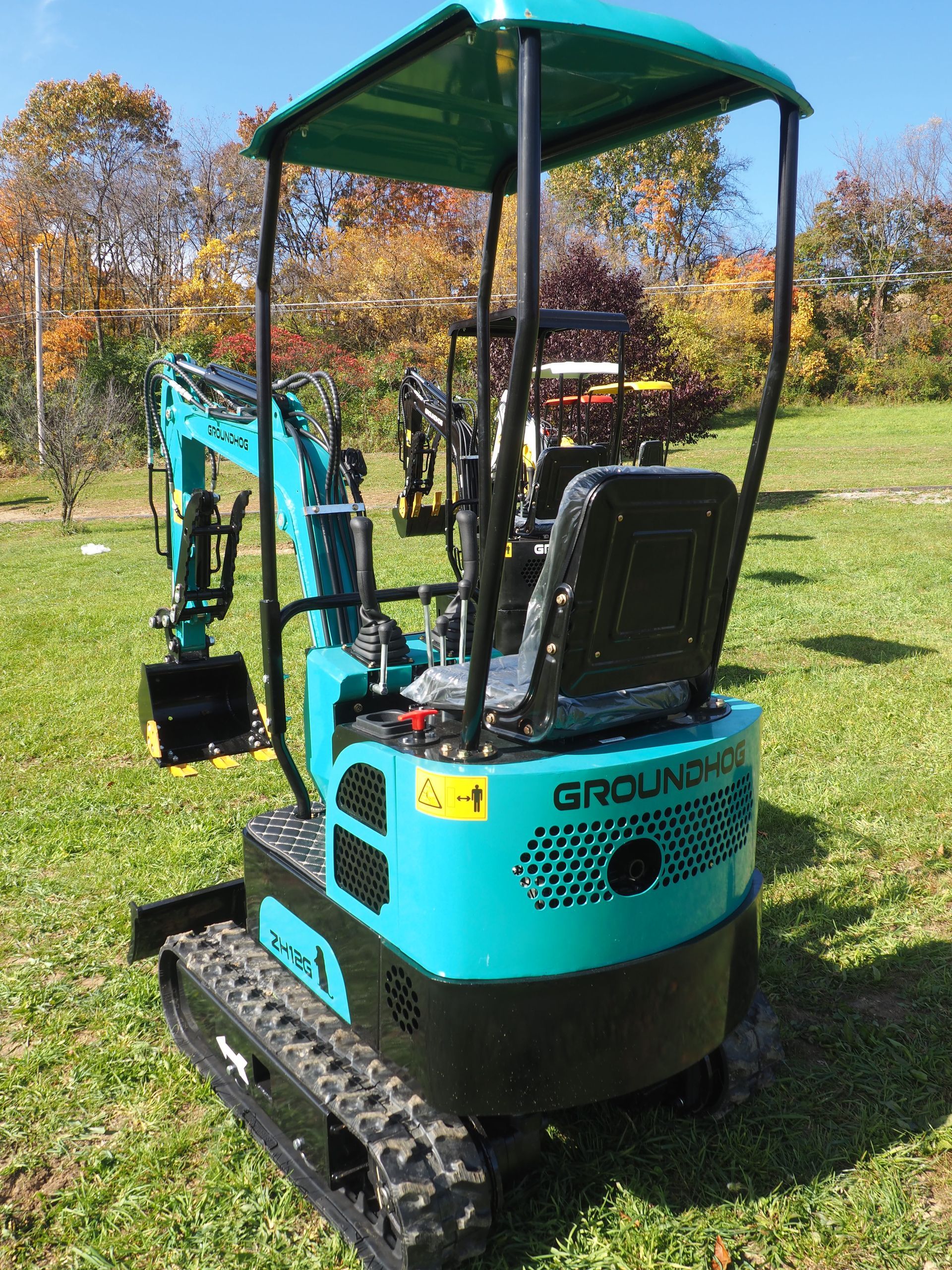 Teal and black mini excavator with protective canopy on green grass; trees in background.