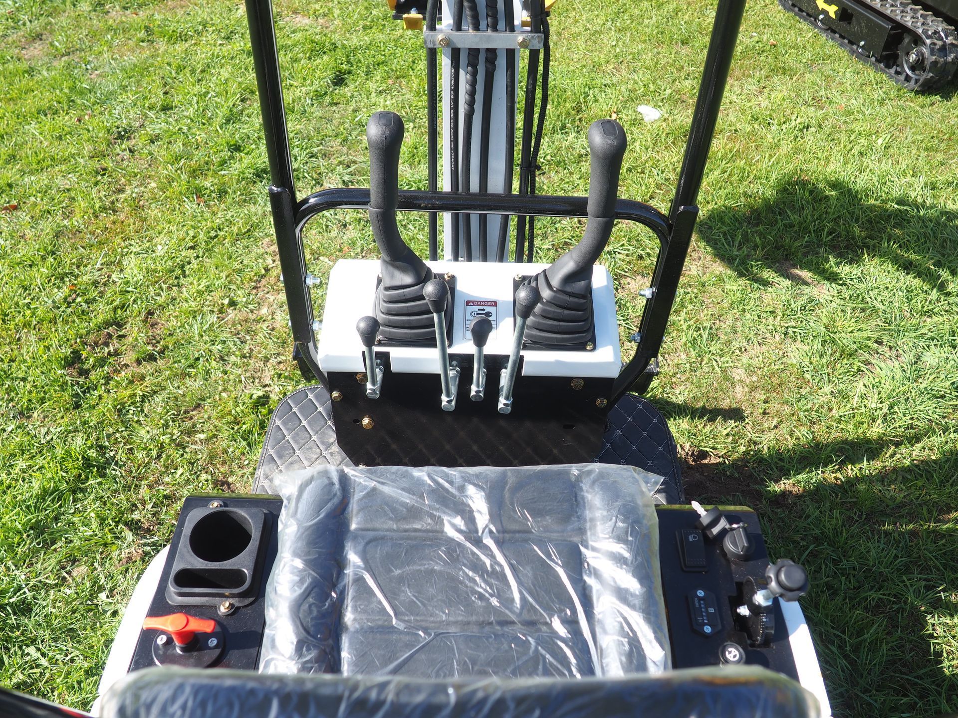Control panel of a small excavator, with levers, a seat covered in plastic, and grass.