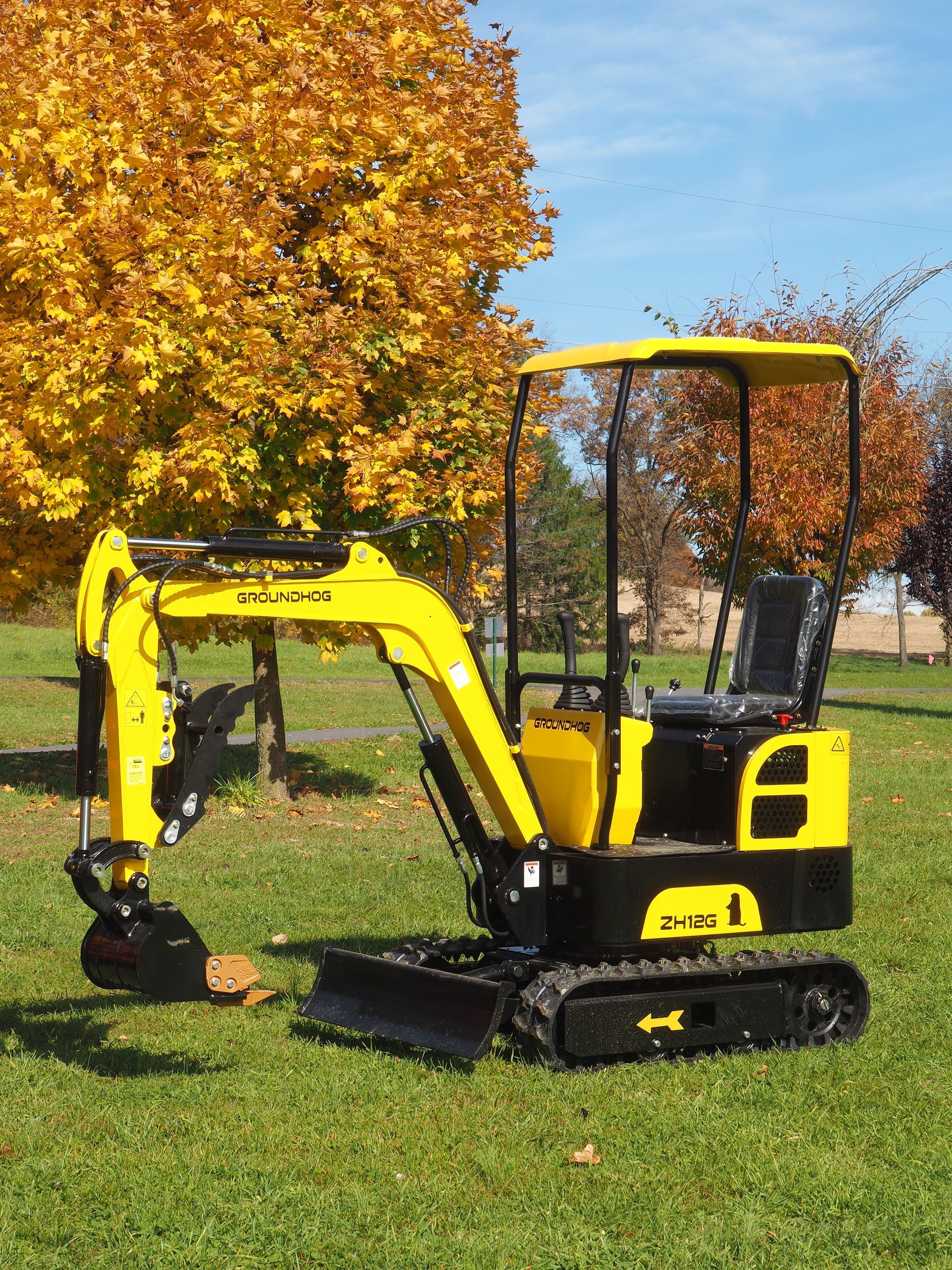 Yellow and black mini excavator on grass in front of a tree with yellow leaves.
