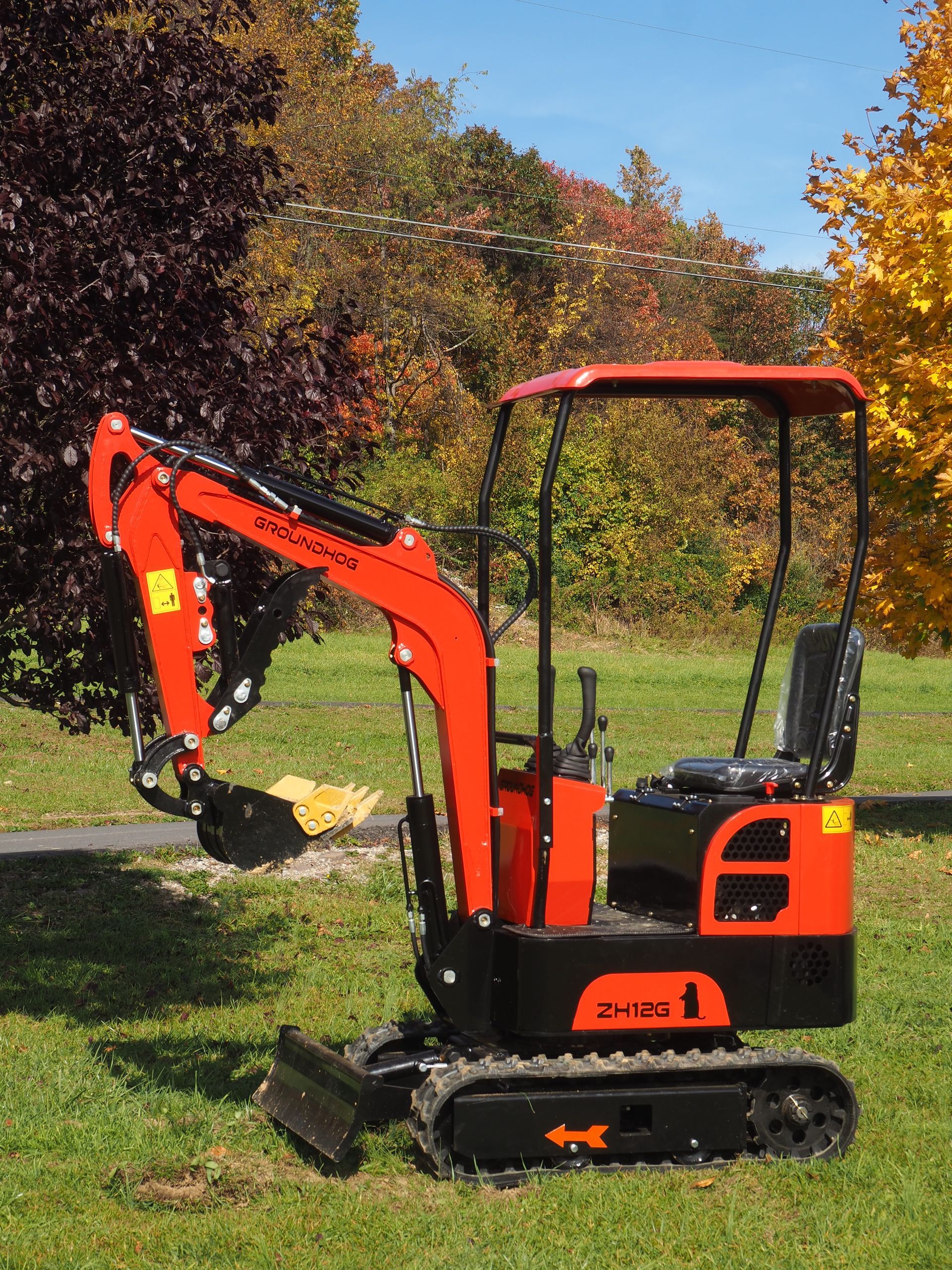 Orange and black mini excavator on grass in front of trees with fall foliage.