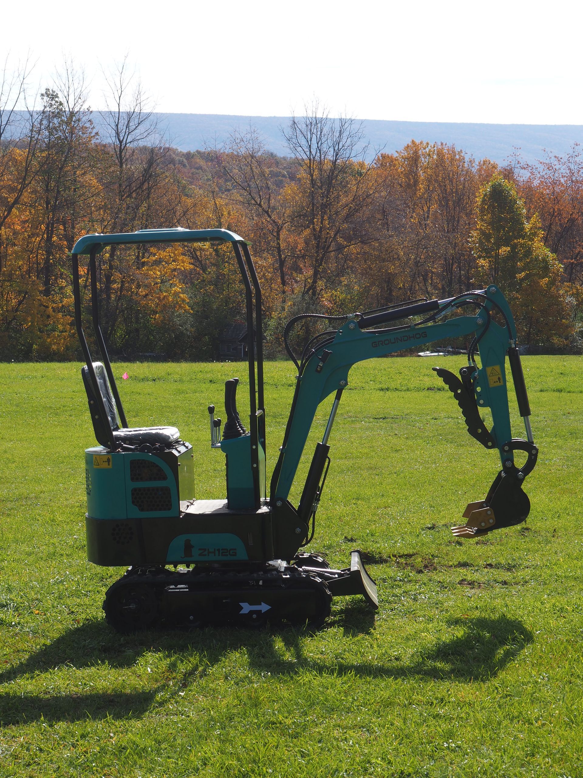 Small teal excavator on grassy lawn with fall foliage background.