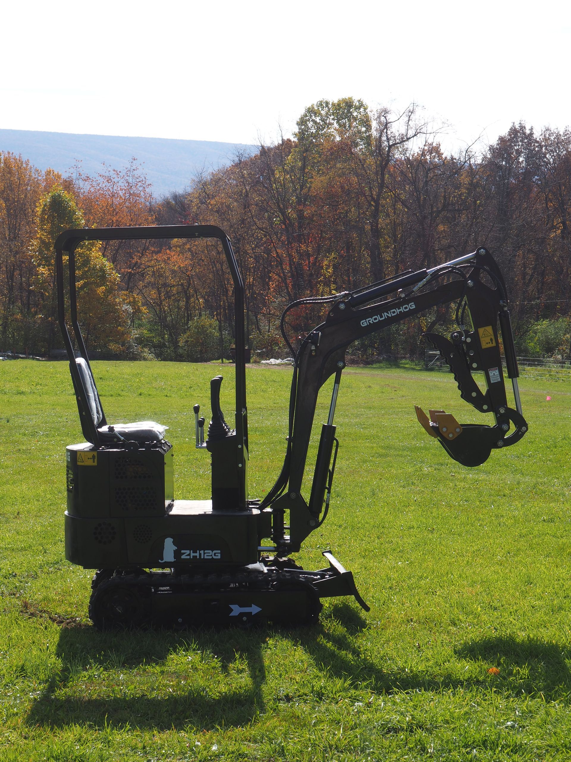 Black mini-excavator on green grass with a tree-filled background.