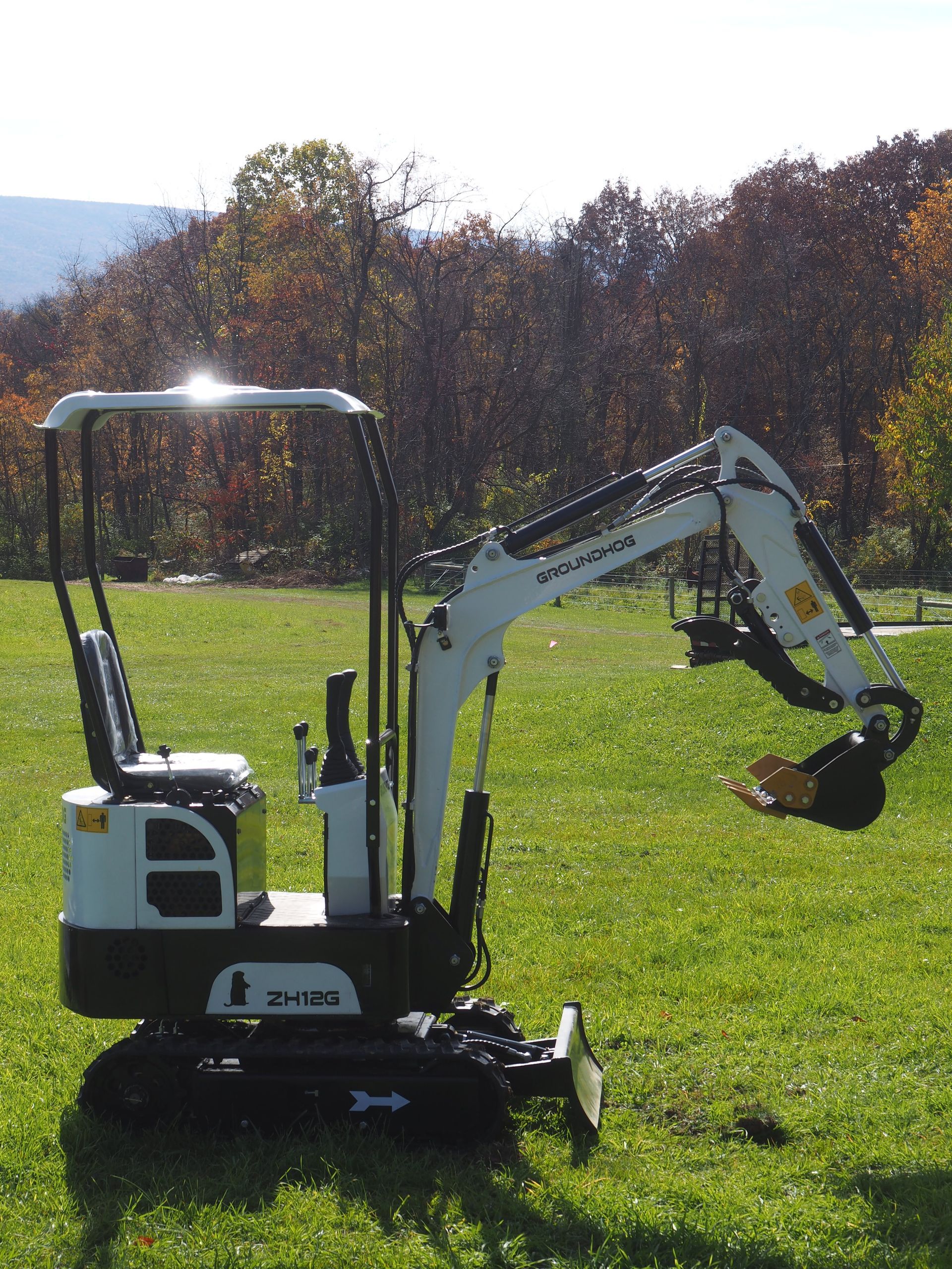 White mini excavator on grass, arm extended, in front of a tree-lined background.