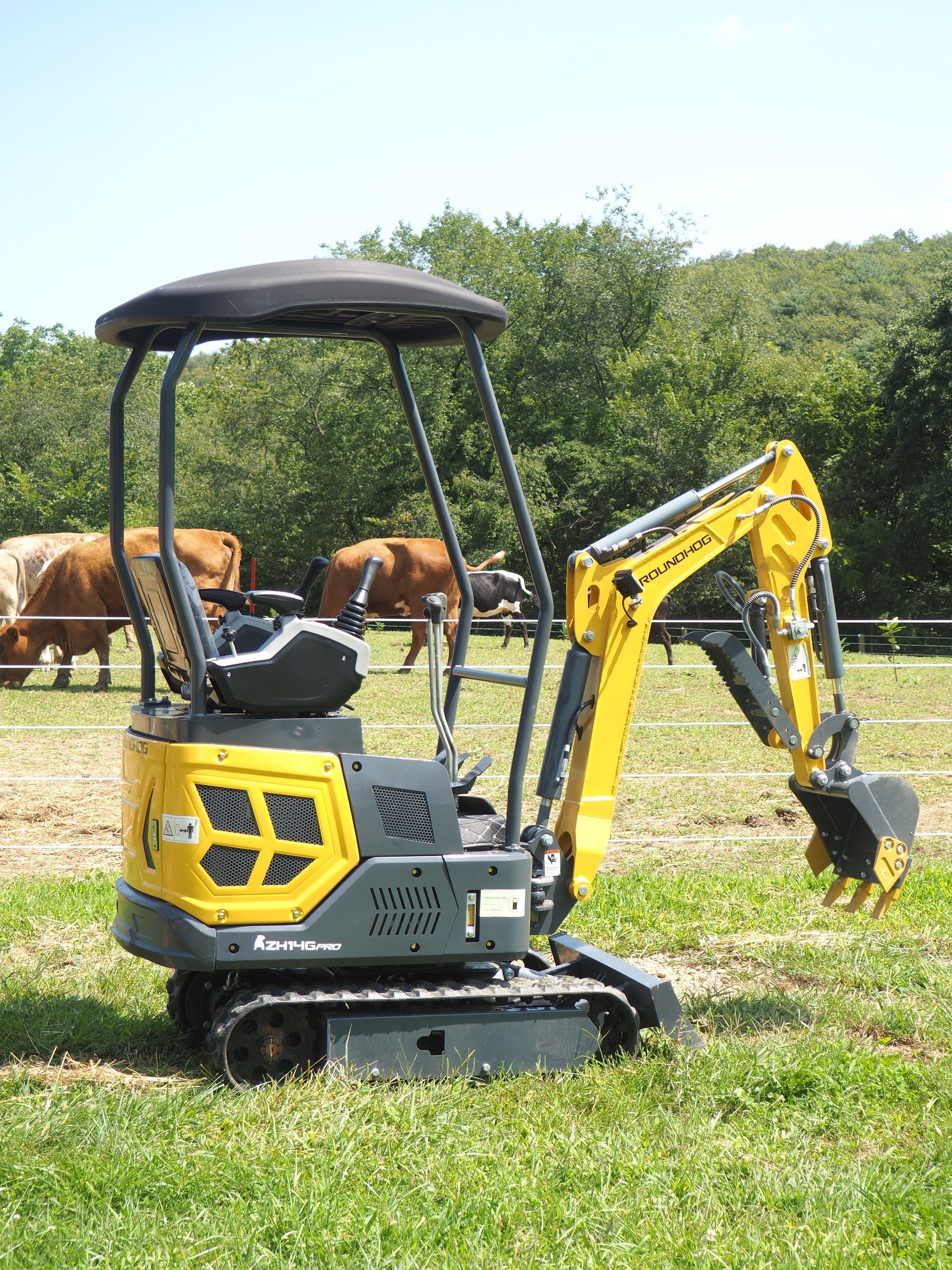Yellow and gray mini excavator on a grassy field with cows in the background.