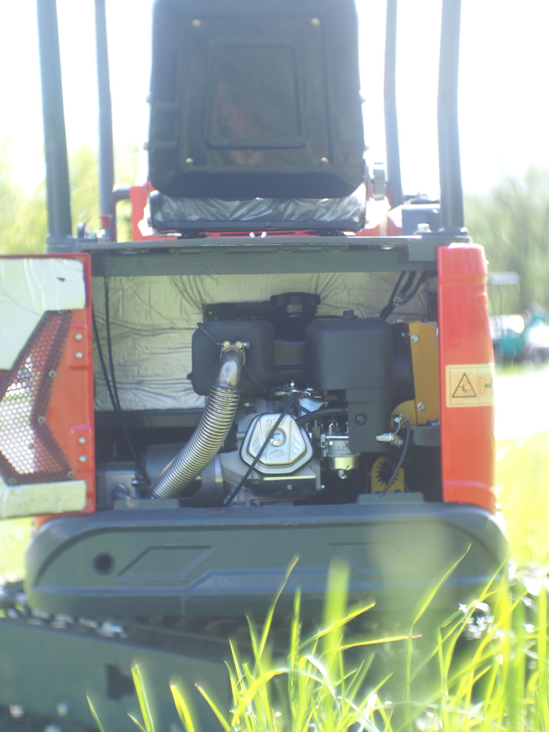 Back view of a small red and black excavator with its engine exposed, outdoors, sunny.
