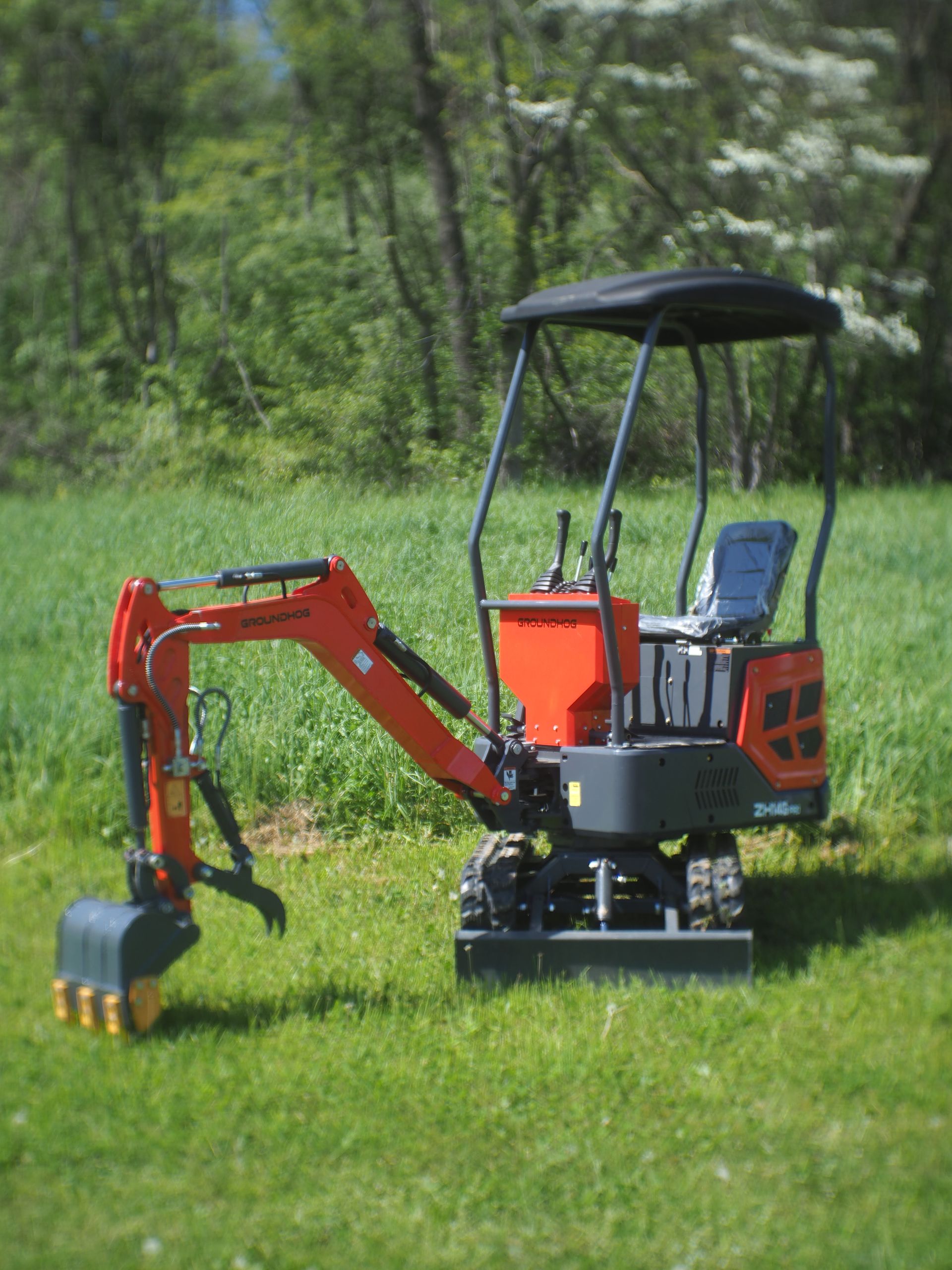 Mini excavator on grass, orange and black, with a tree-lined background.