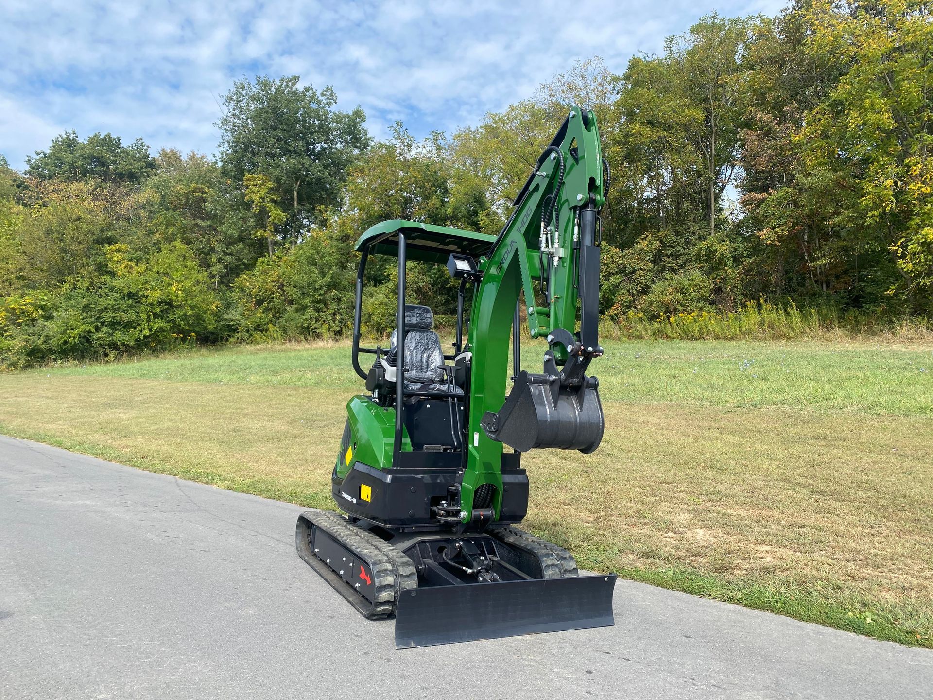Green mini excavator on asphalt road in a field, trees in the background.