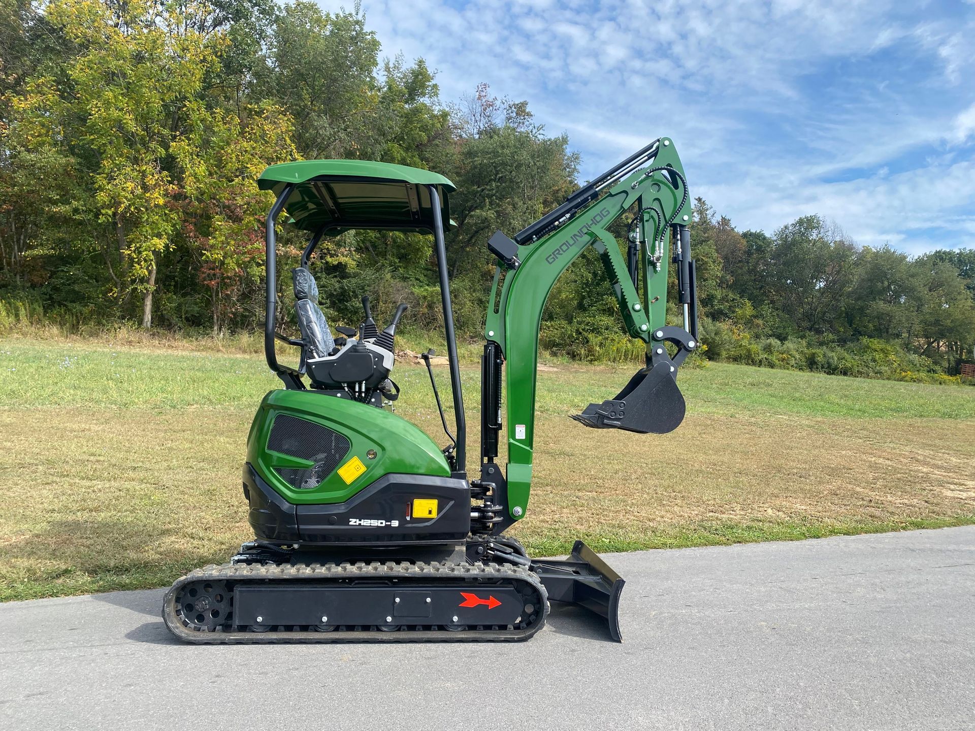 Green mini excavator on a paved area, in front of a grassy field and trees.
