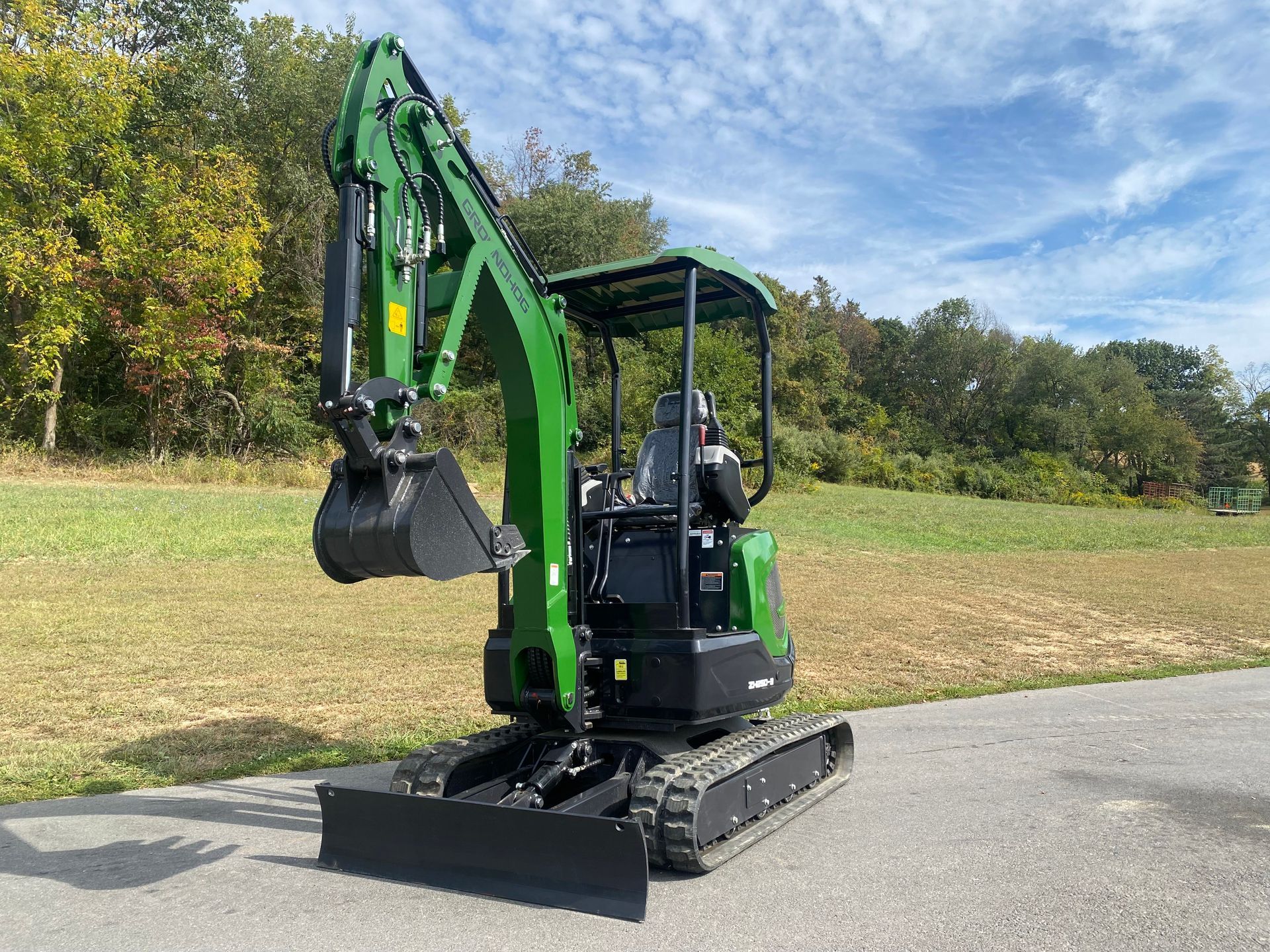Green mini excavator on a paved surface, with a grassy field and trees in the background.