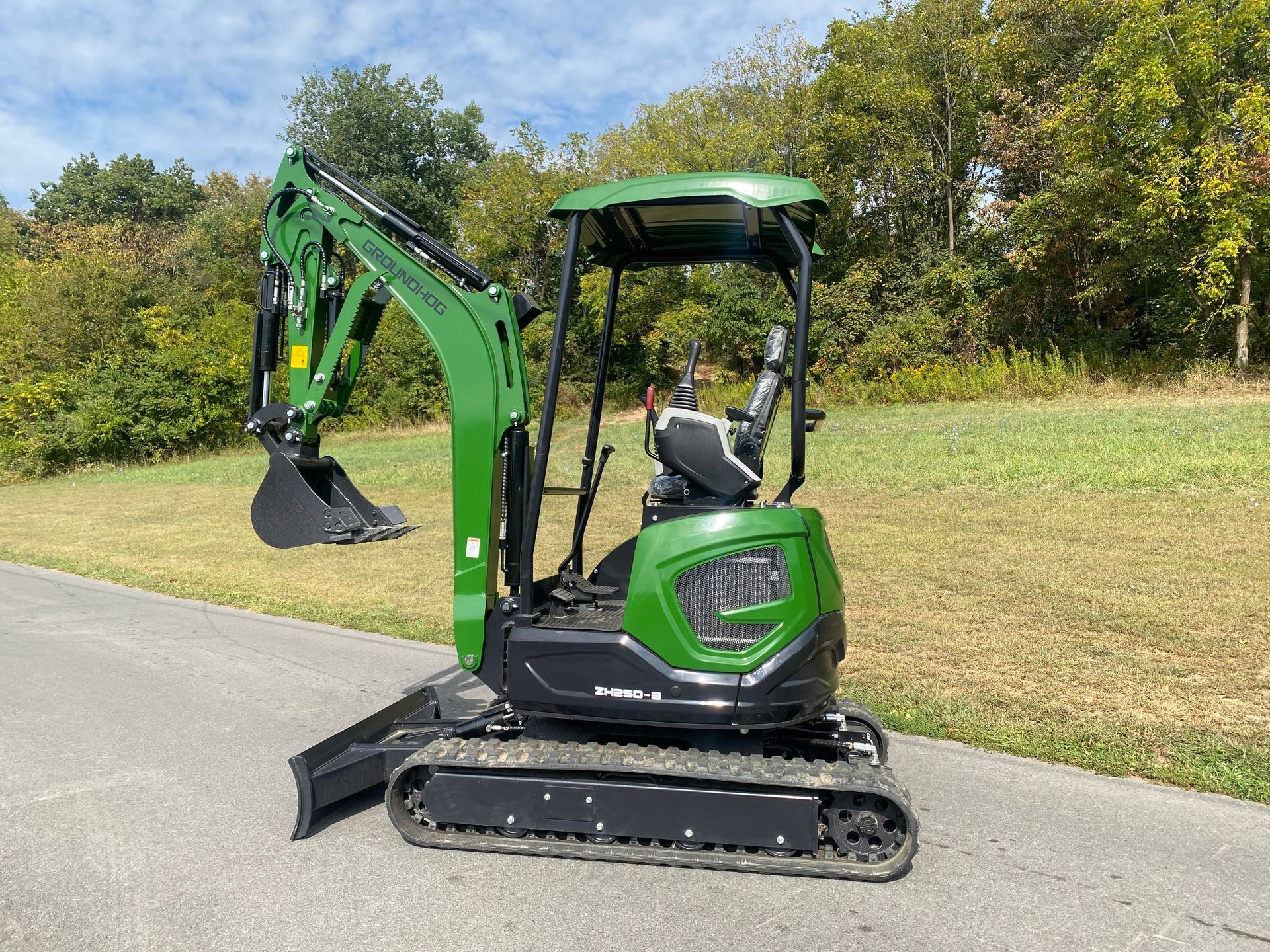 Green mini excavator parked on asphalt with a wooded backdrop.