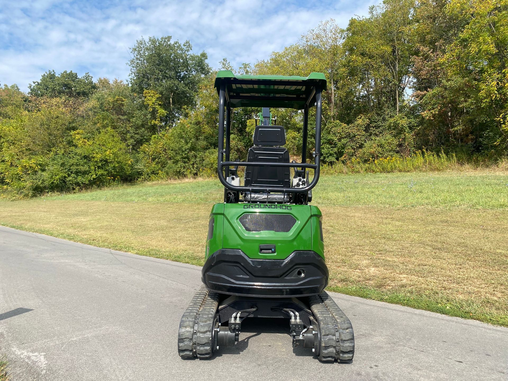 Green and black mini excavator on a paved surface, with a grassy field and trees in the background.