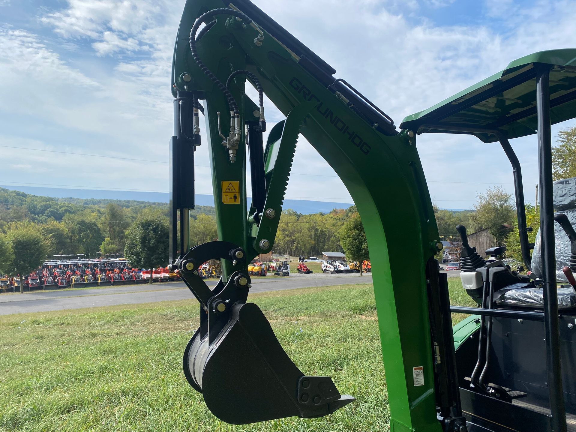 Green excavator arm and bucket, outdoors with field, crowd, and mountains in background.