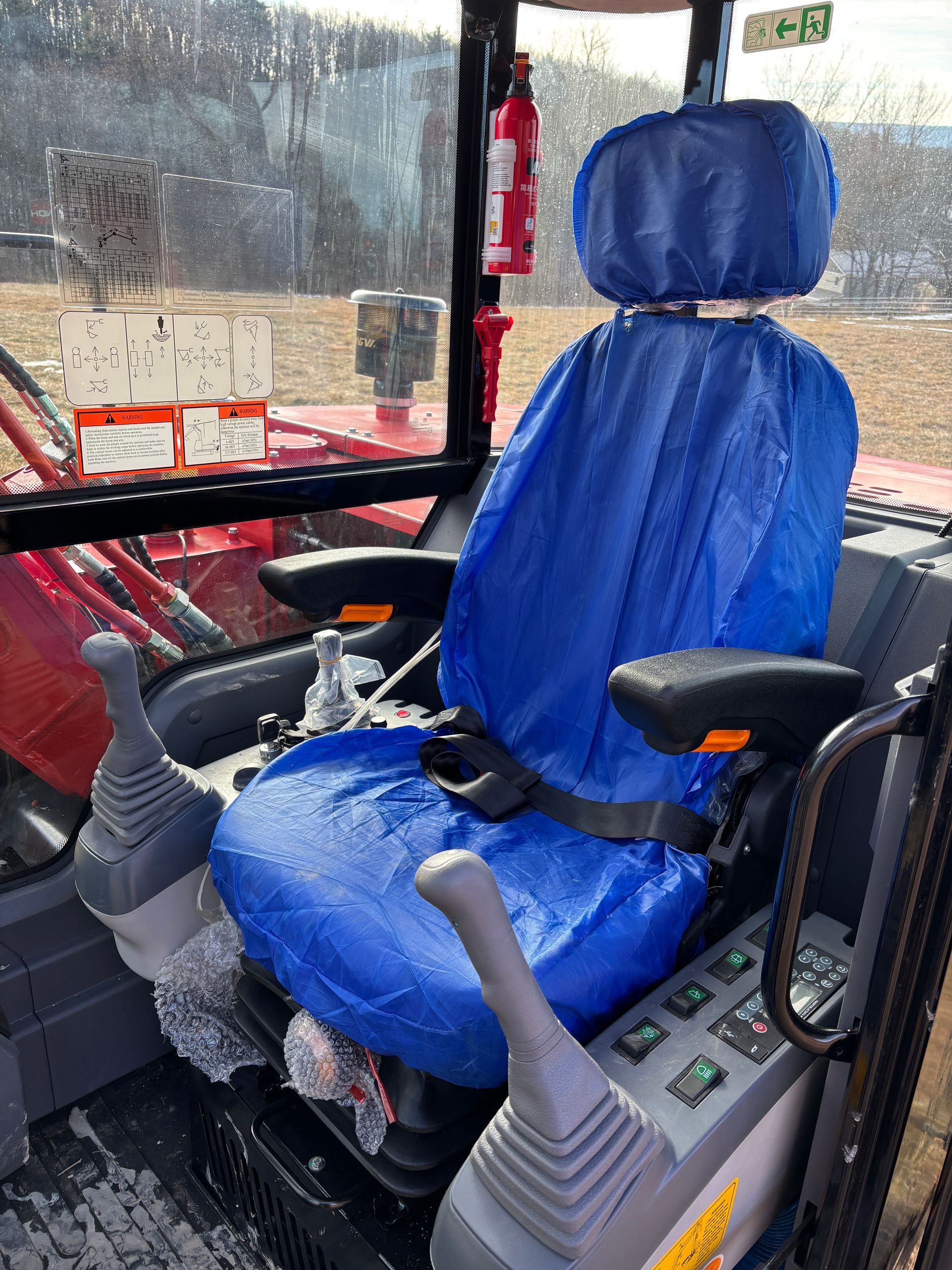 Interior of an excavator cab with a blue seat cover, control sticks, and a fire extinguisher.