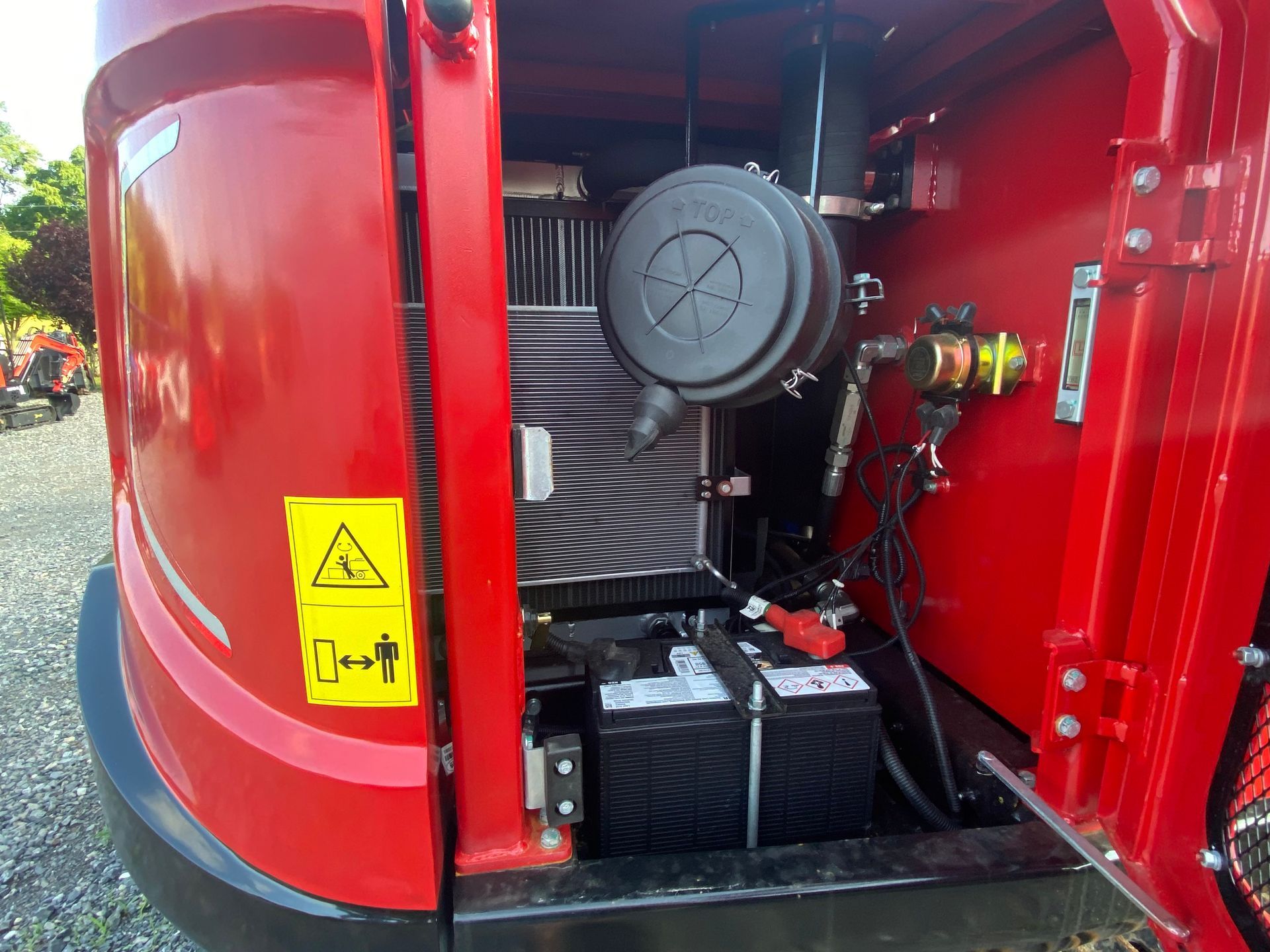 Red machine's open compartment reveals engine components, including batteries, air filter, and warning label.