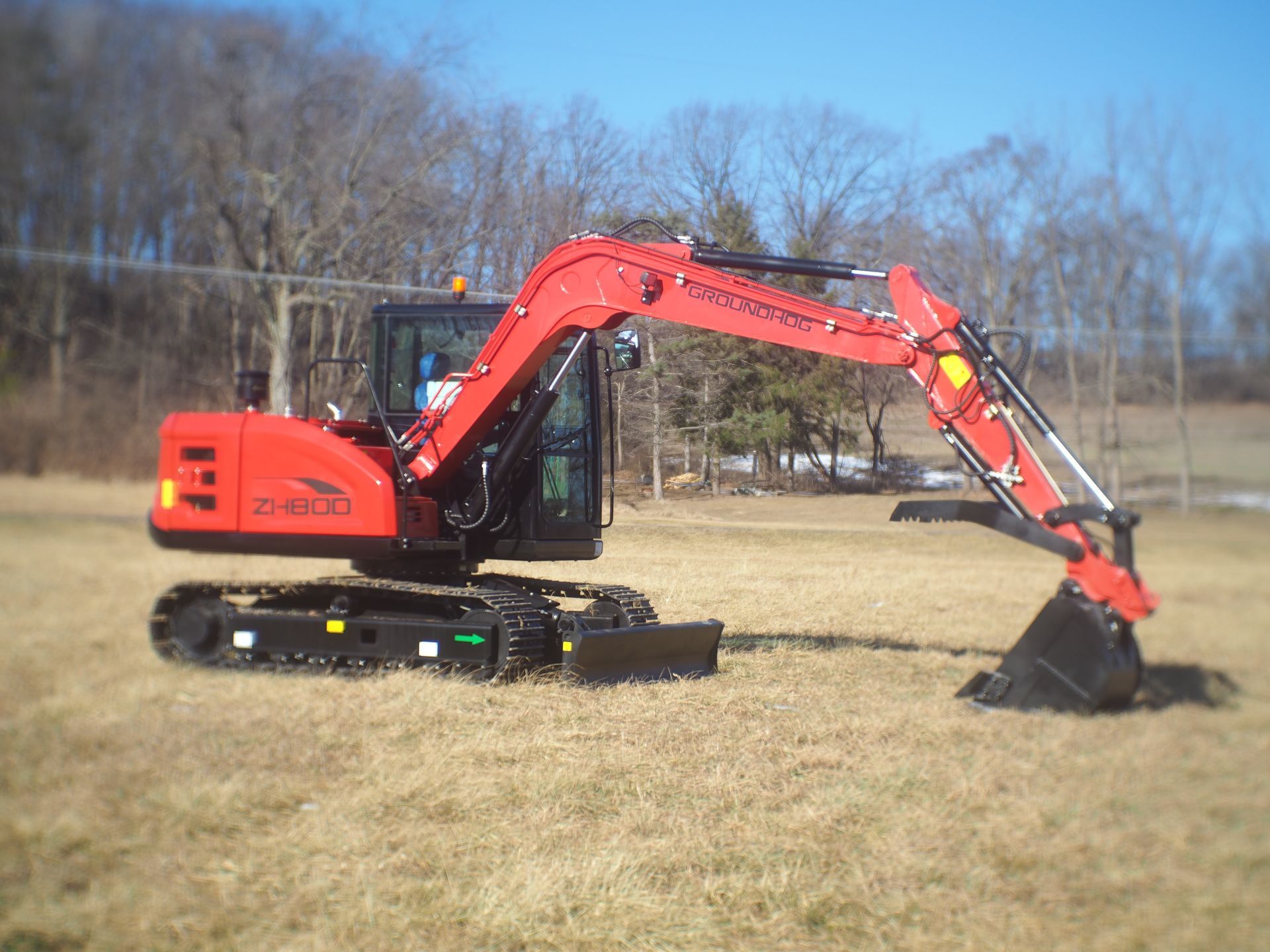 Red mini excavator on a grassy field; in front of leafless trees.