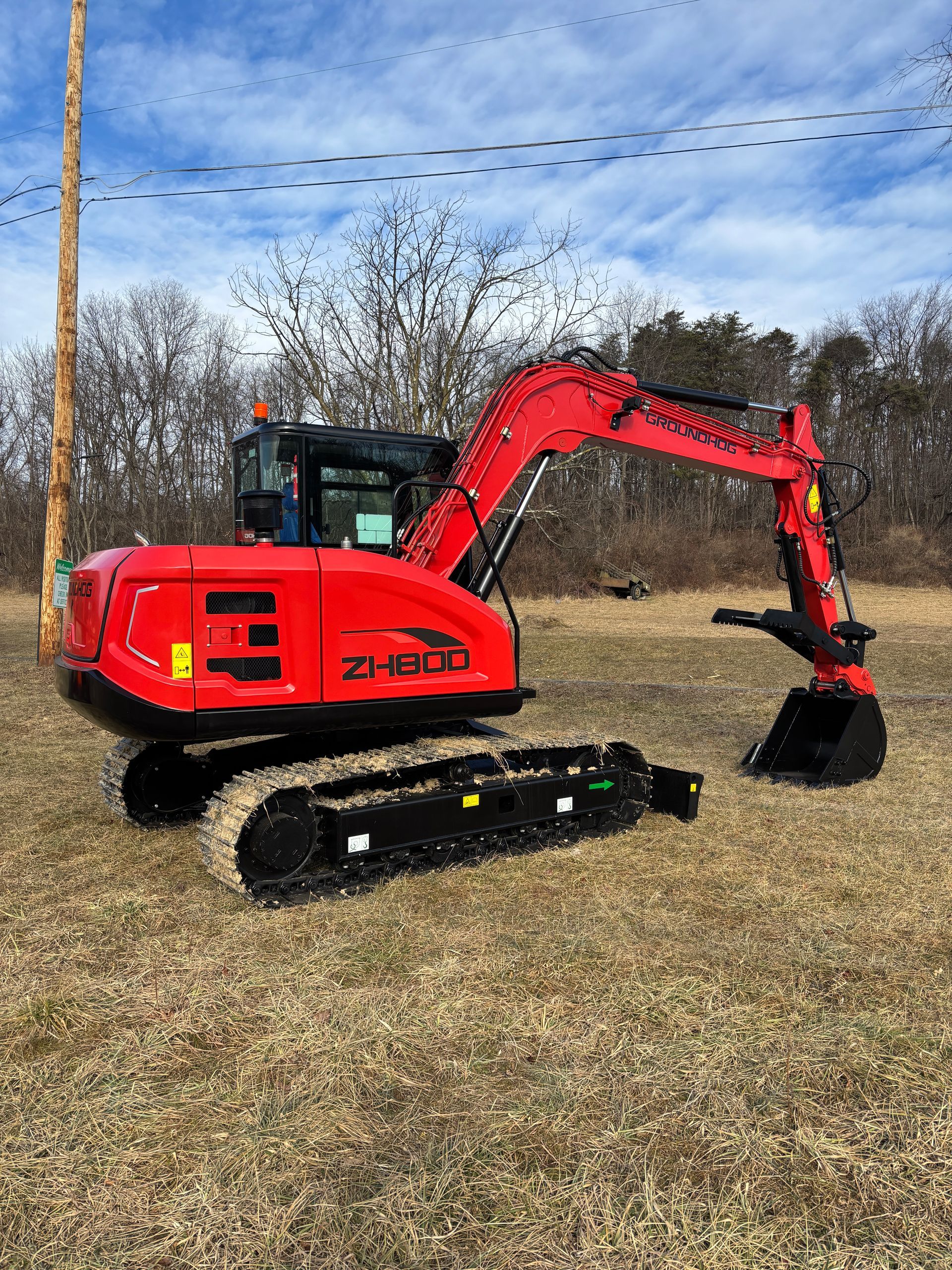 Red excavator on a grassy field under a blue sky with power lines and trees.