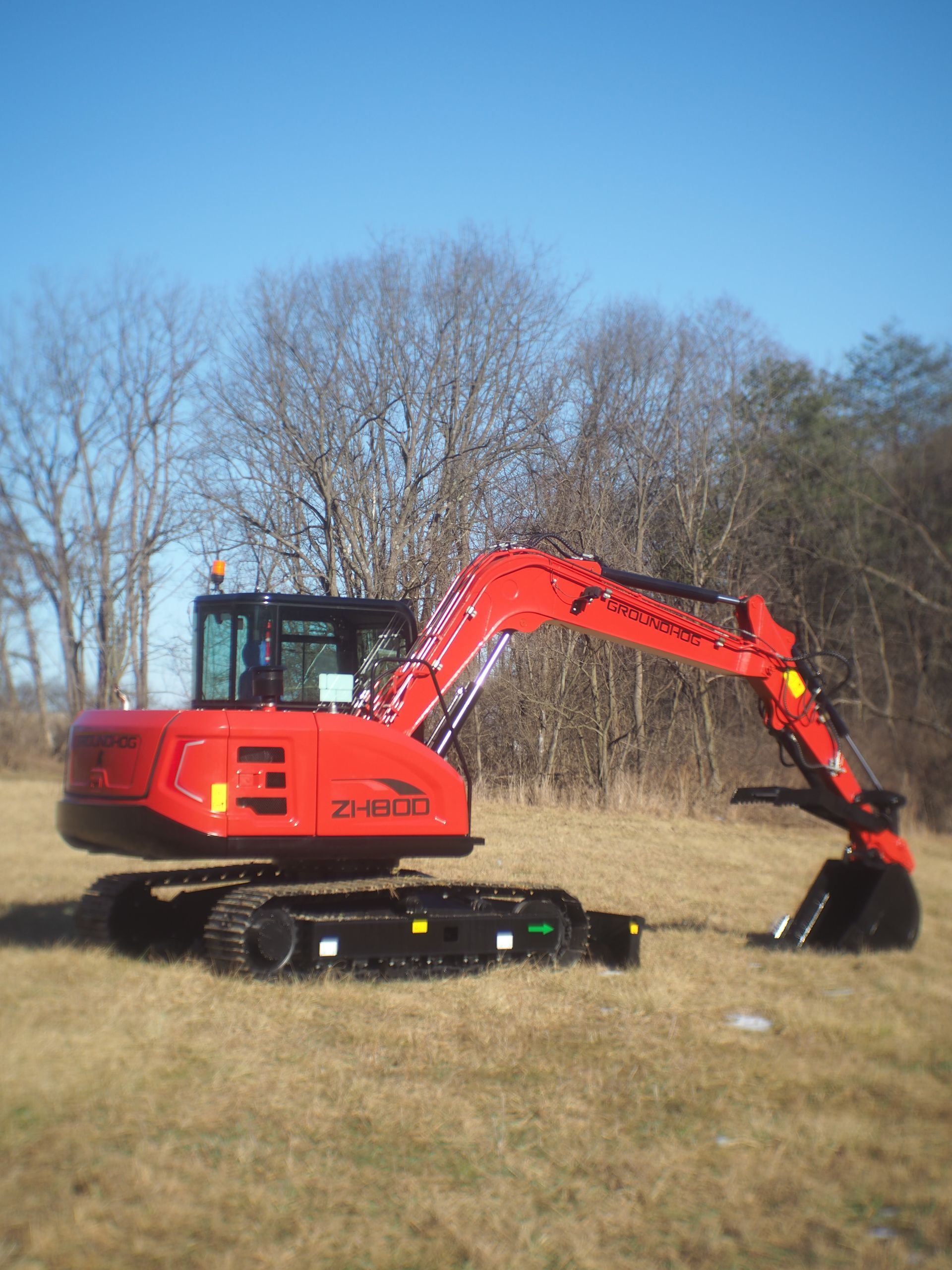 Red excavator on tracks in a grassy field with bare trees and a blue sky.