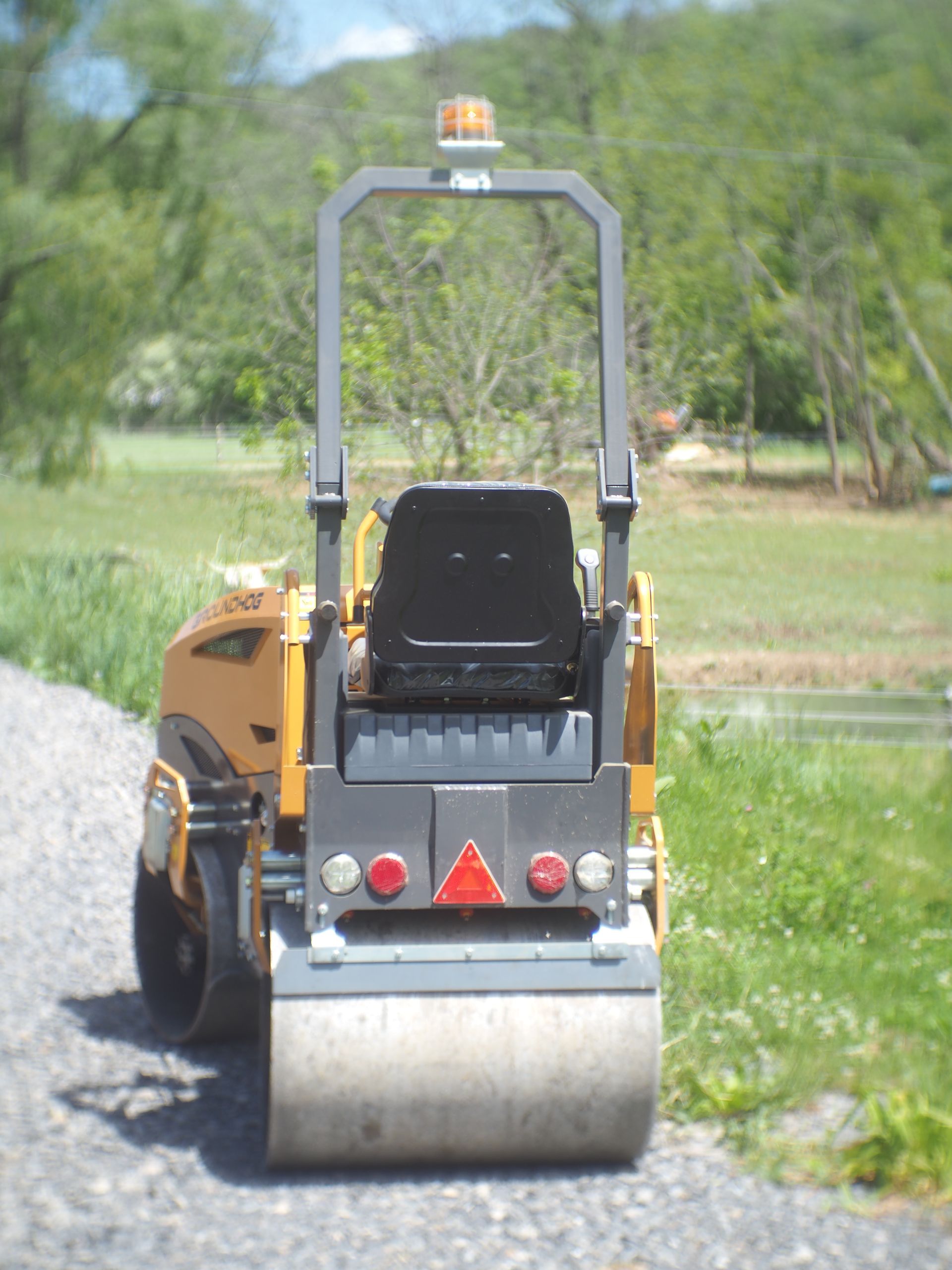 Yellow road roller on a gravel path, back view, with a safety cage, lights, and a seat; countryside background.