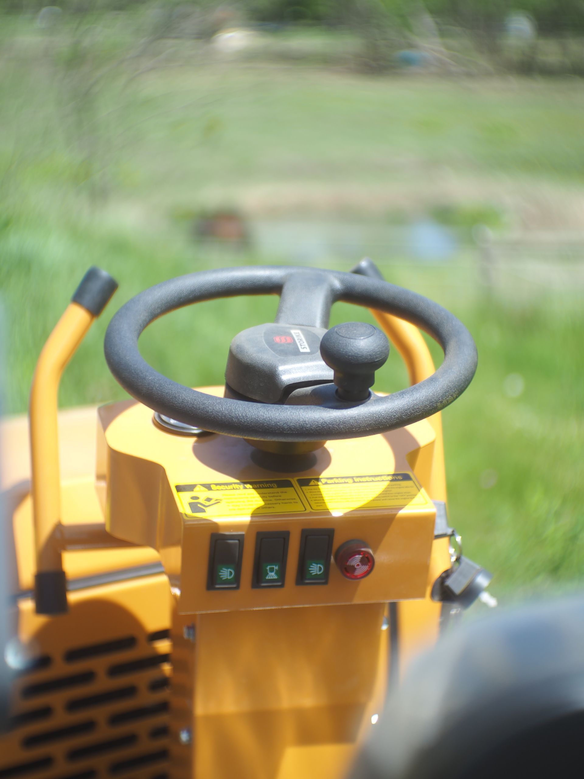 Yellow and black riding lawnmower steering wheel and controls against a blurred green background.