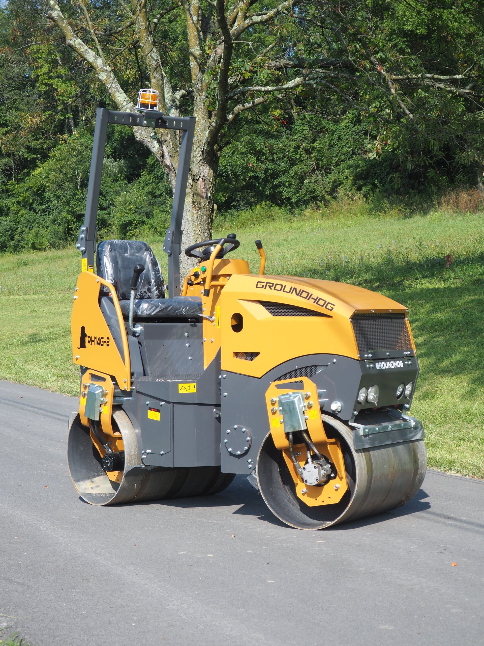 Yellow and gray road roller compacting asphalt on a paved road, set against a green grassy background.