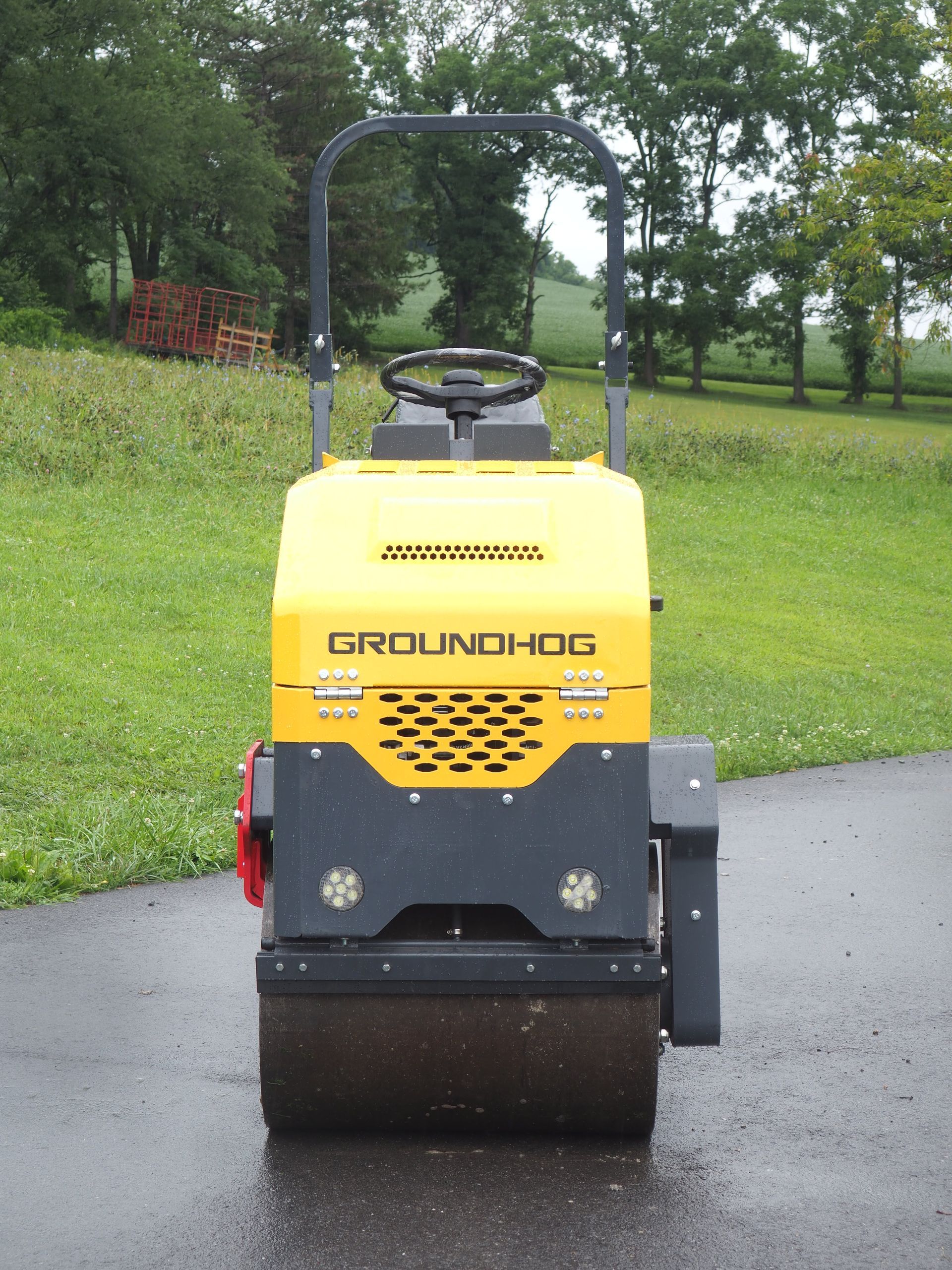 Yellow and gray Groundhog road roller on a paved surface, with a green lawn and trees in the background.