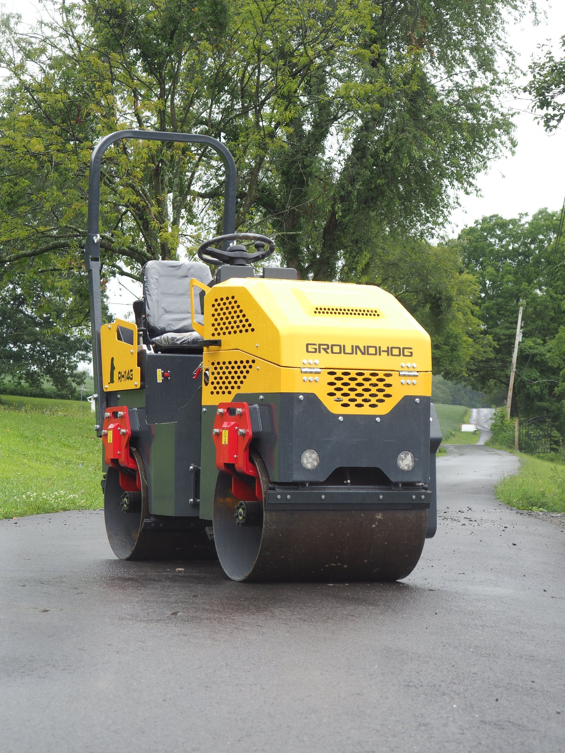 Yellow and gray road roller compacting asphalt on a road, set against a backdrop of trees.