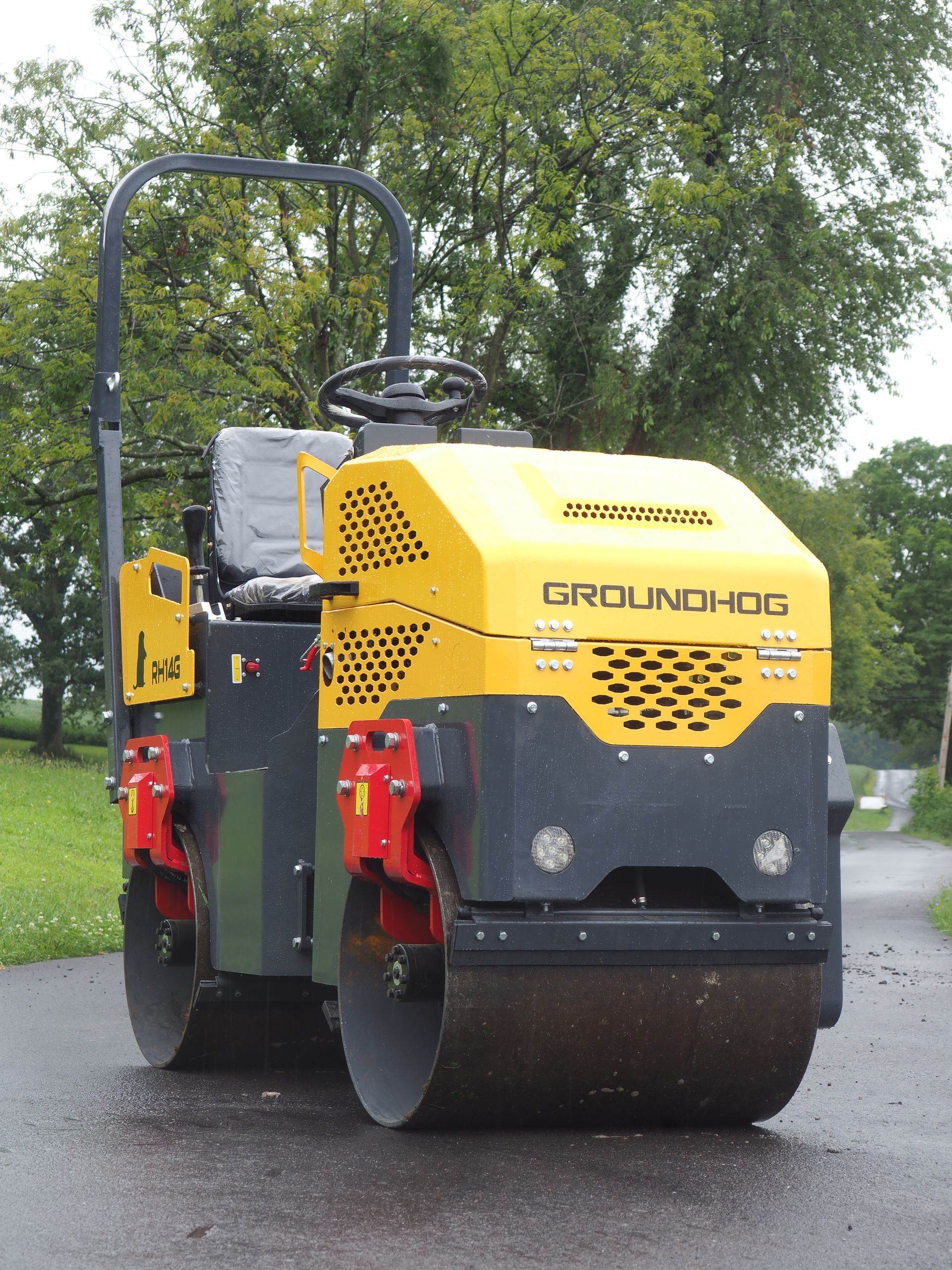 Yellow and black Groundhog road roller on asphalt in front of trees.