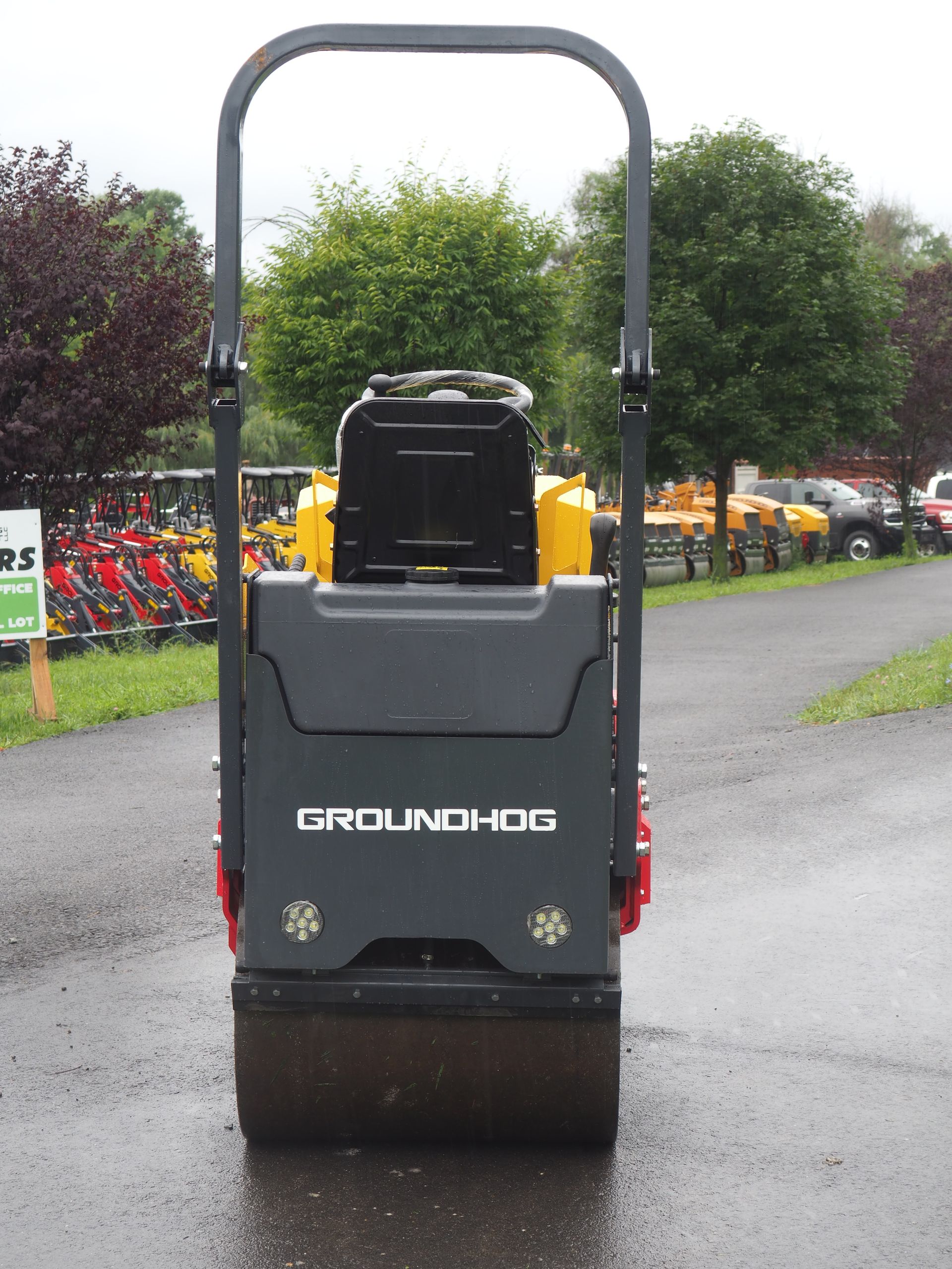 Rear view of a Groundhog plate compactor; dark grey with a yellow engine, handle and roller outdoors.