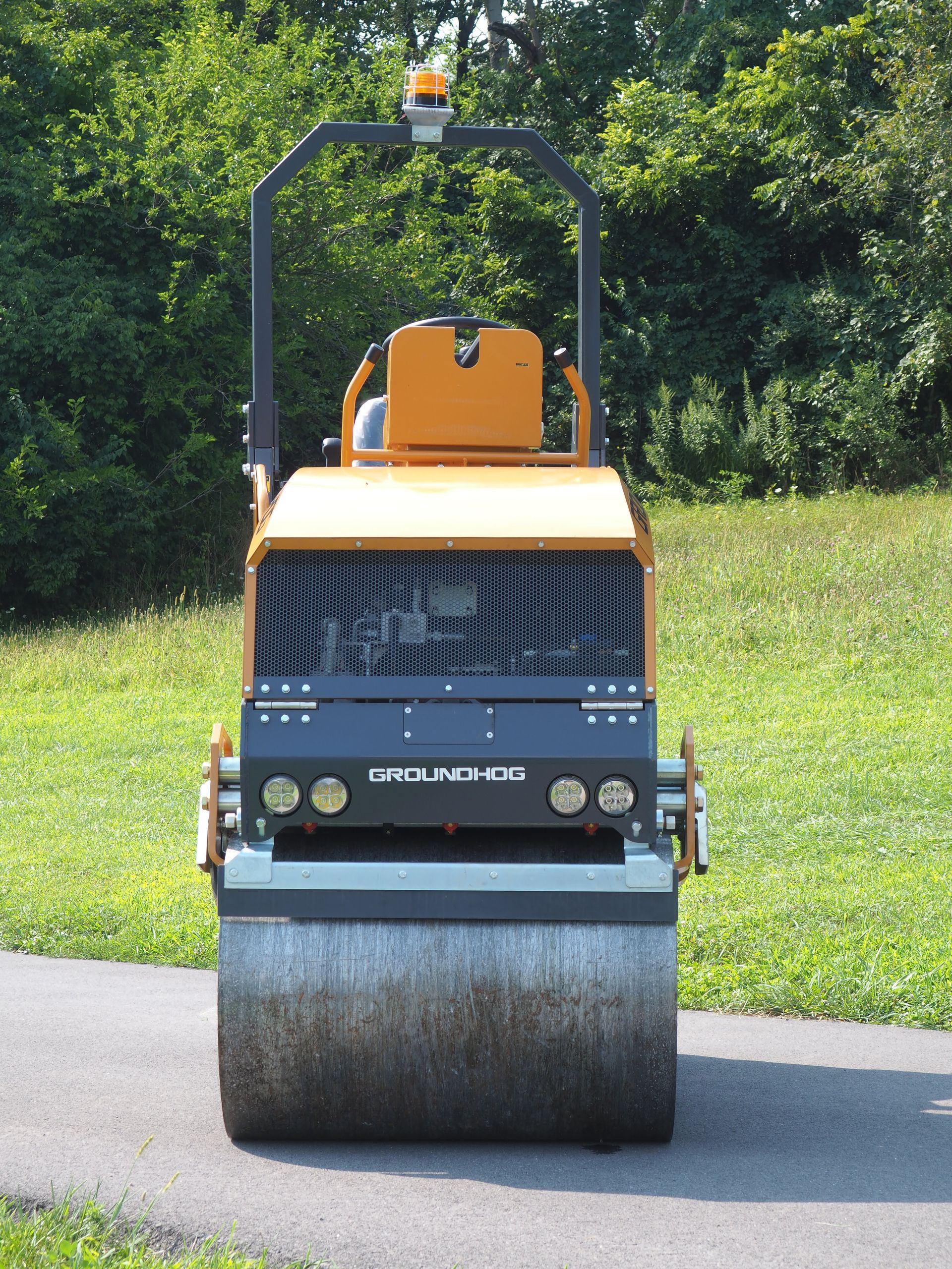 Yellow and black road roller on pavement with green grass and trees in the background.