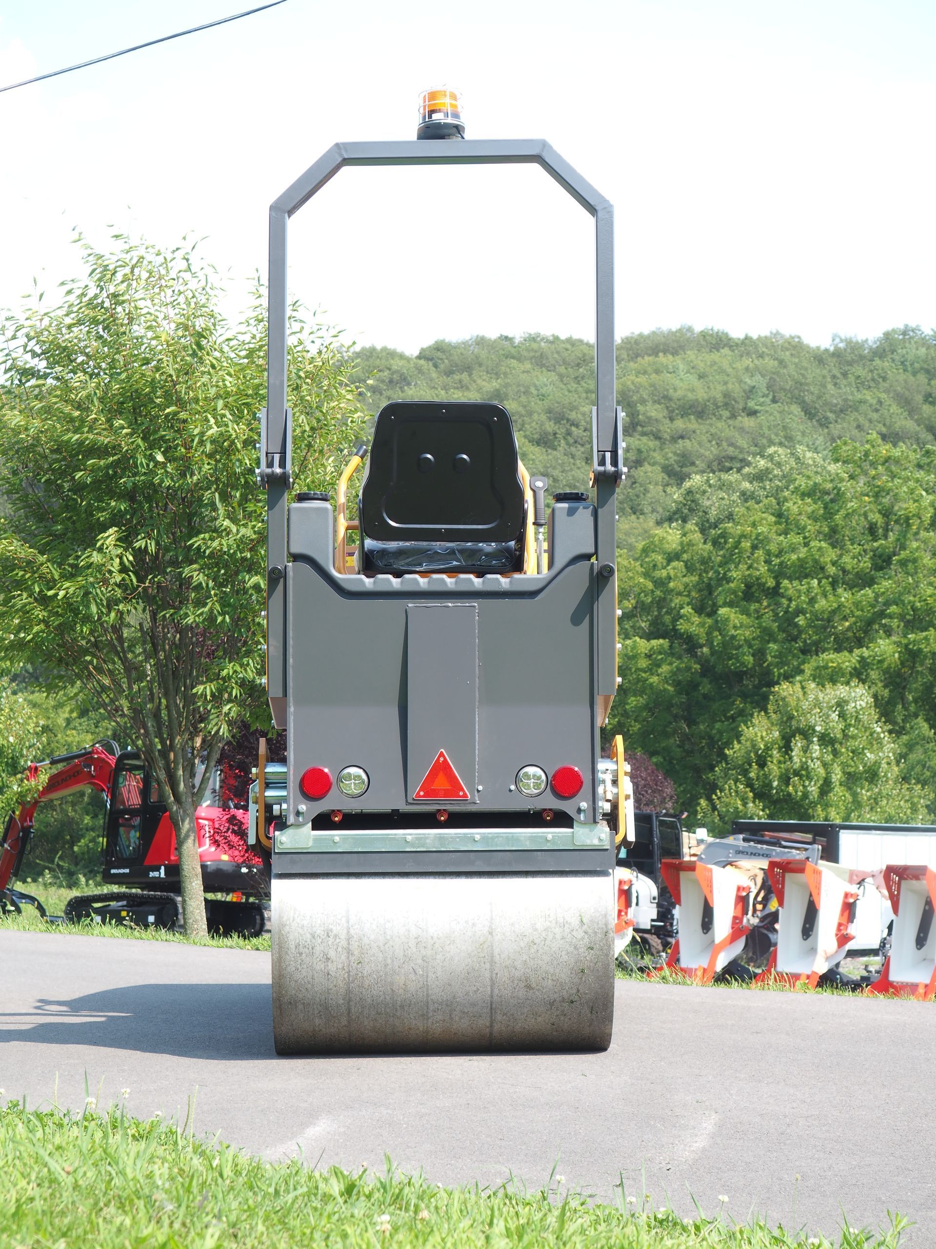 Road roller on paved surface, rear view, gray with visible drum and safety lights, greenery in the background.