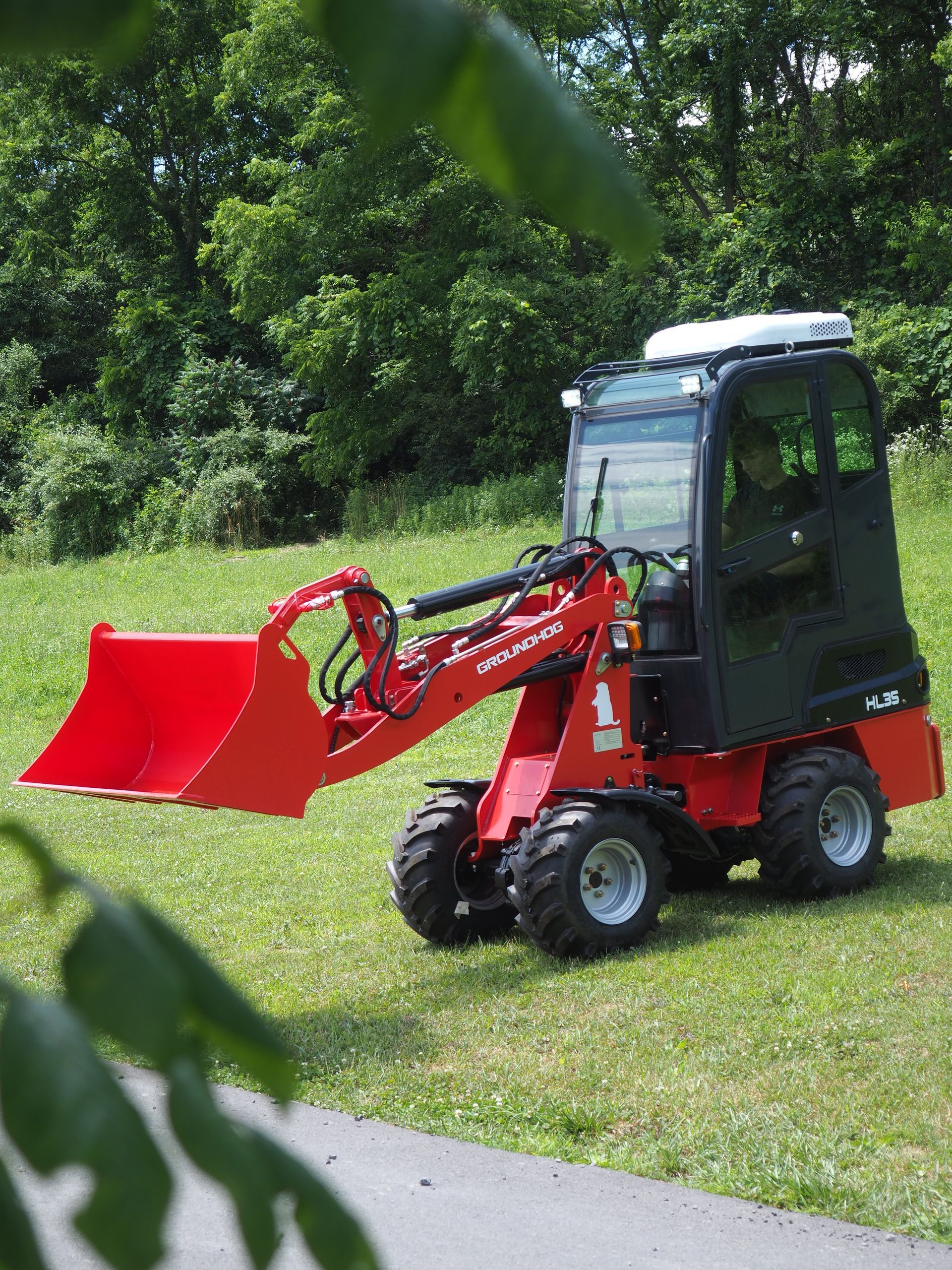 Red compact loader on grass, near trees. Black cab, bucket extended.