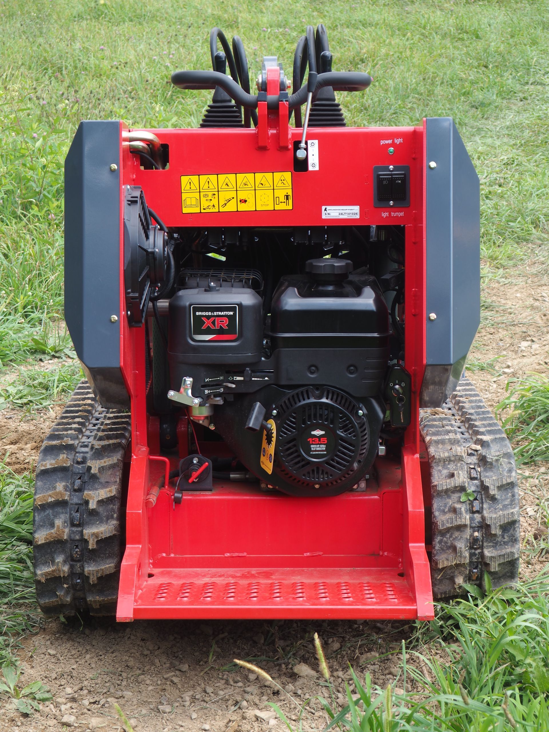 Red and black mini-skid steer with tracks, engine bay open, outdoors on grass.
