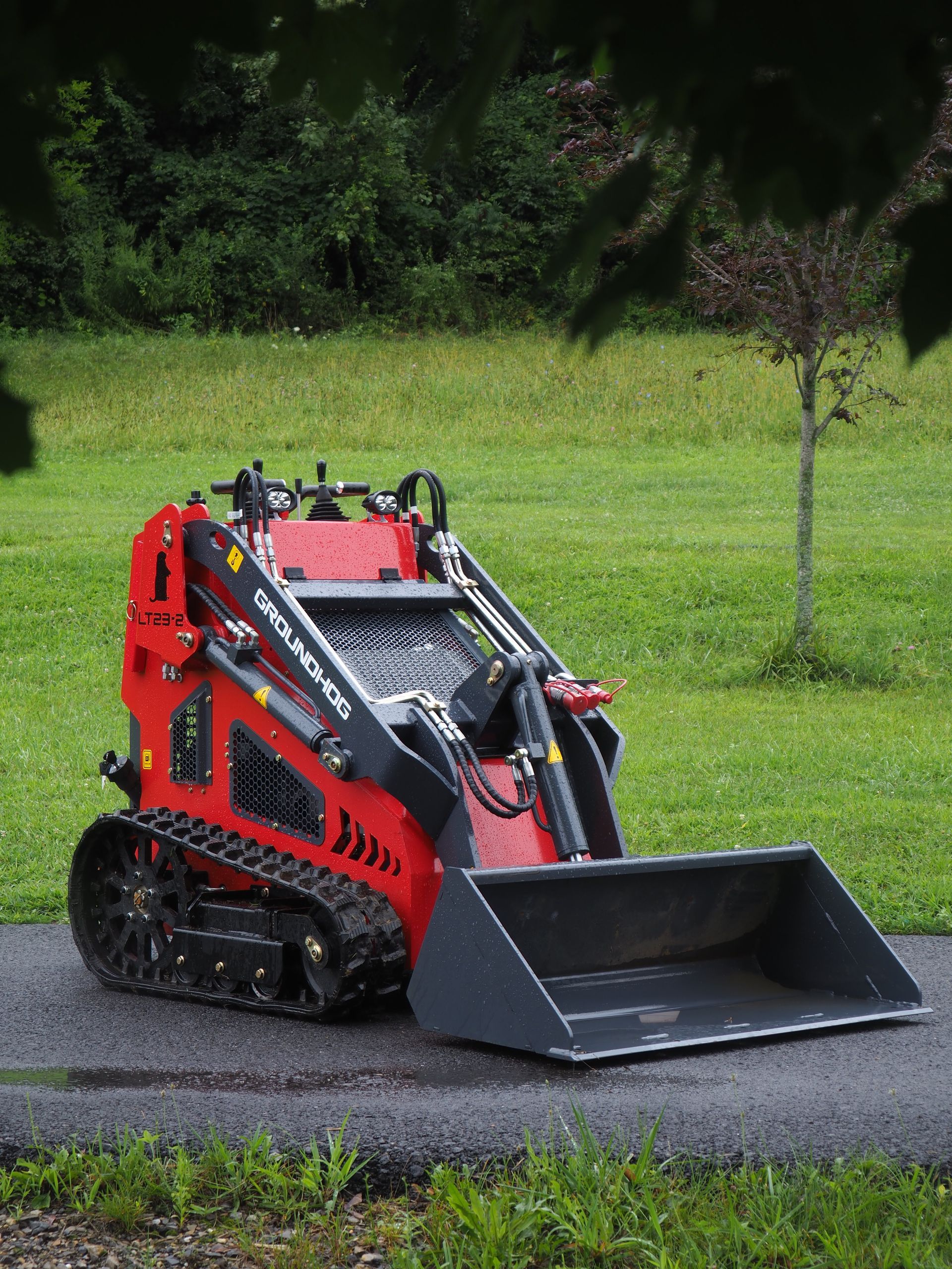 Red mini track loader with a bucket on asphalt, green grass and trees in the background.