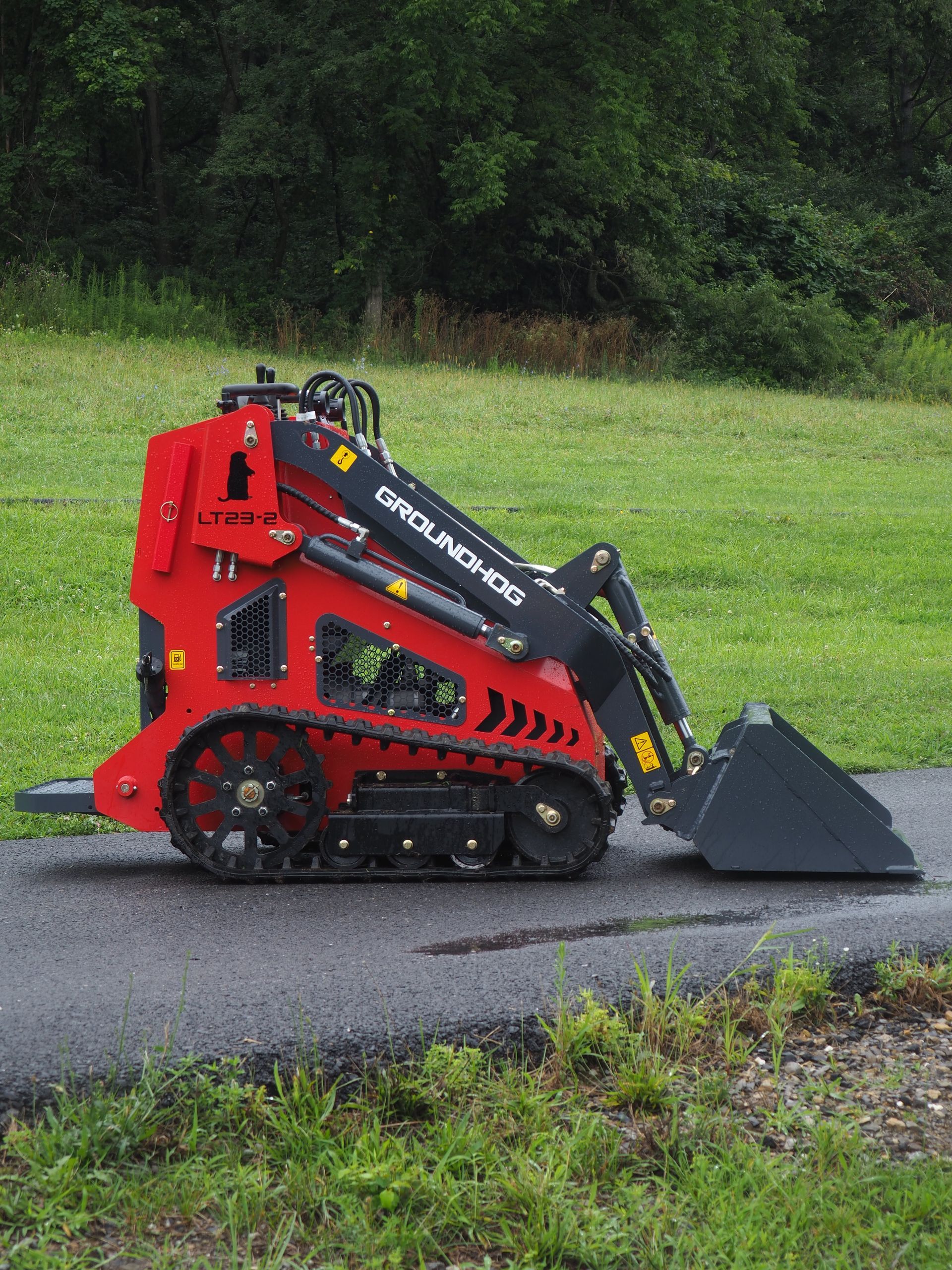 Red mini-skid steer with black tracks and bucket on asphalt, in a grassy outdoor setting.