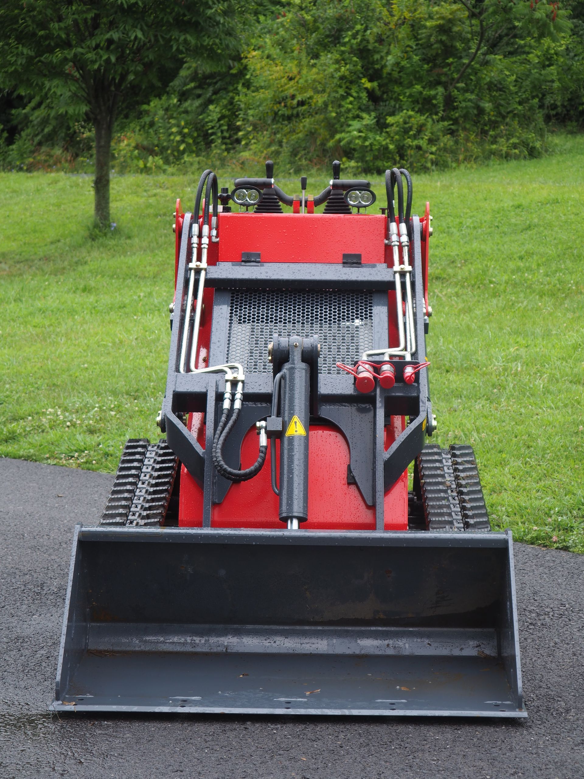 Red and black mini track loader on asphalt, bucket down, outdoors.