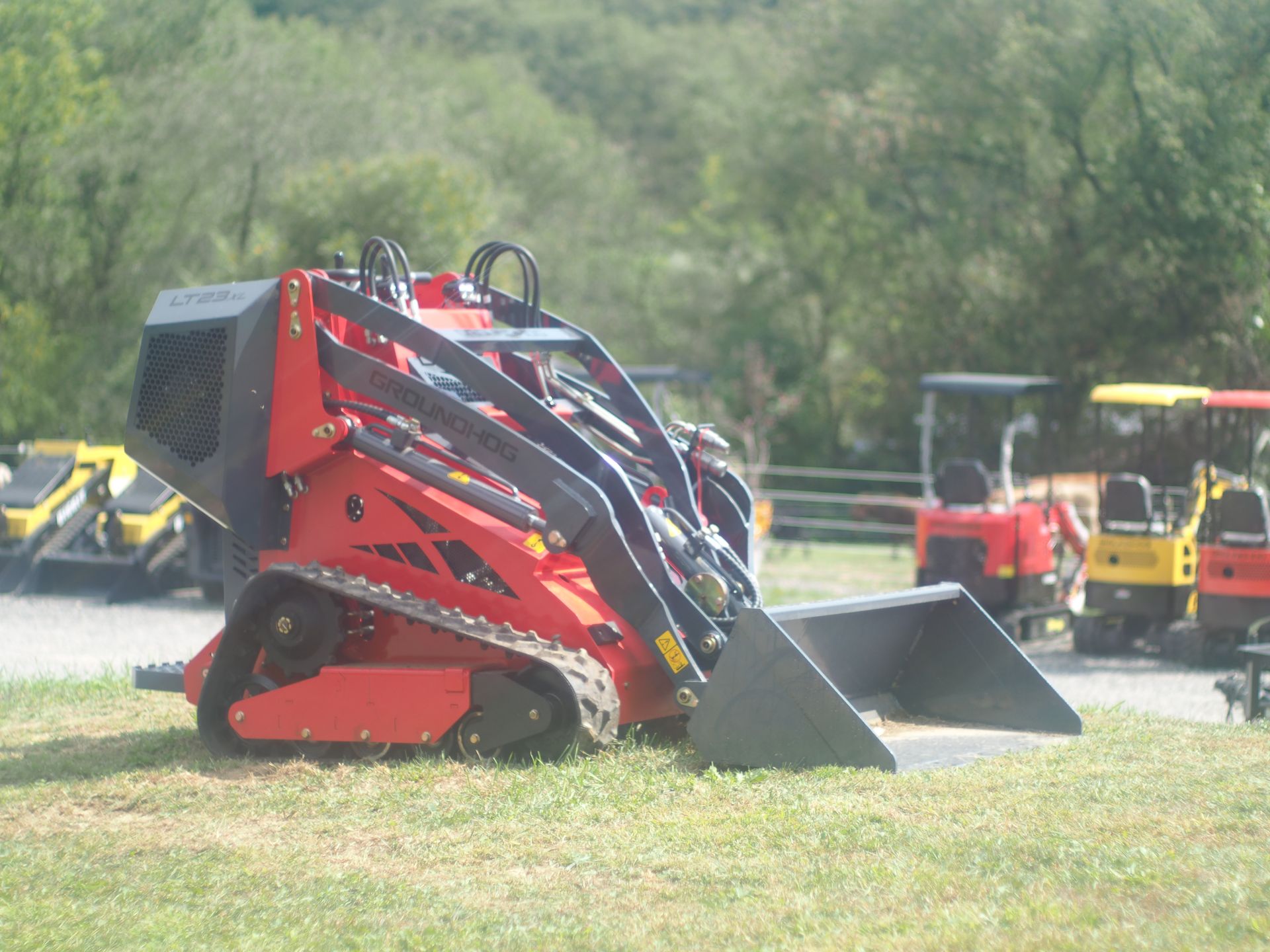 Red compact track loader with a bucket, in a grassy area with other machines.