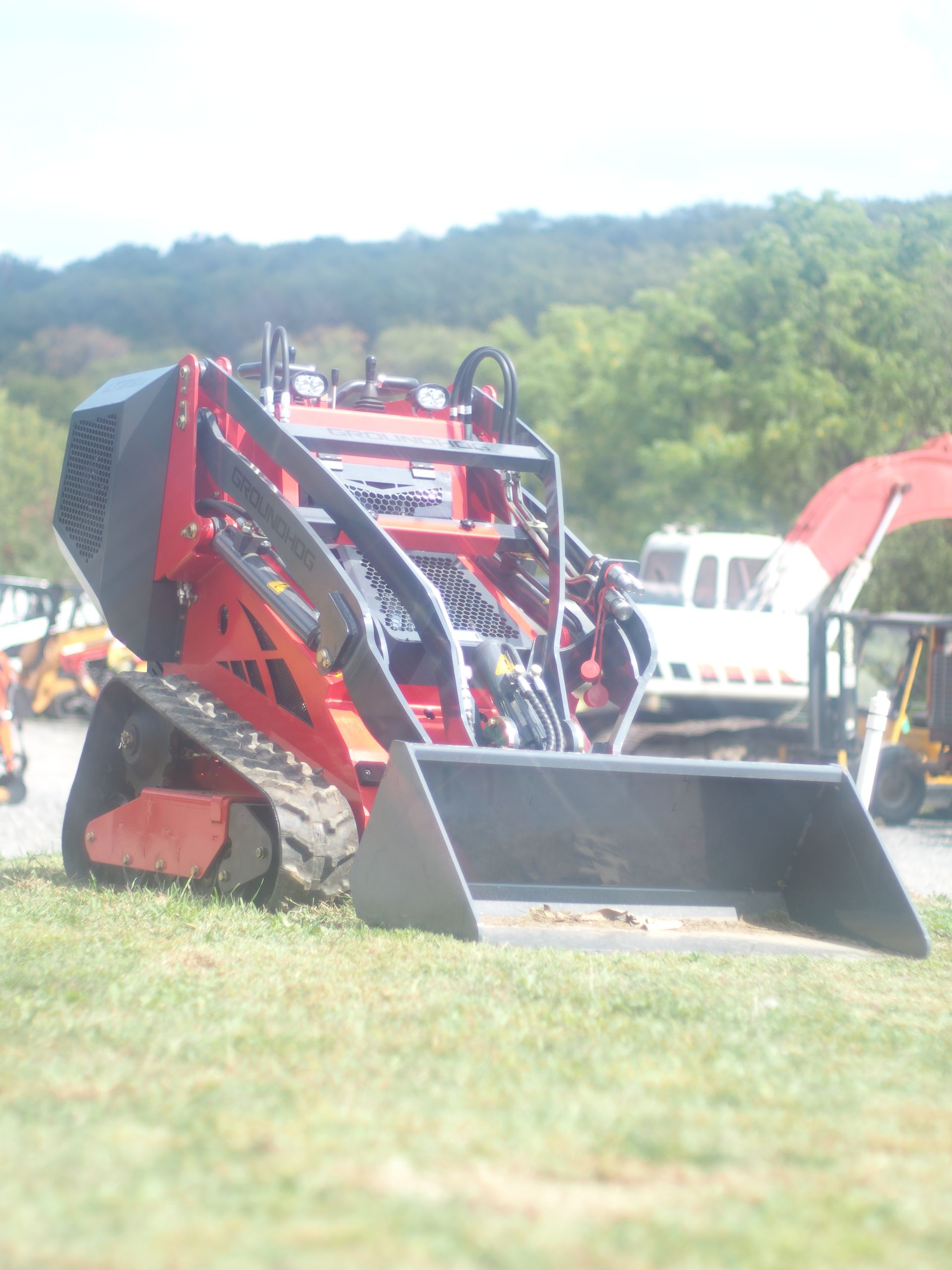 Red and black tracked mini-skid steer loader with a bucket, parked on grass. Trees in the background.